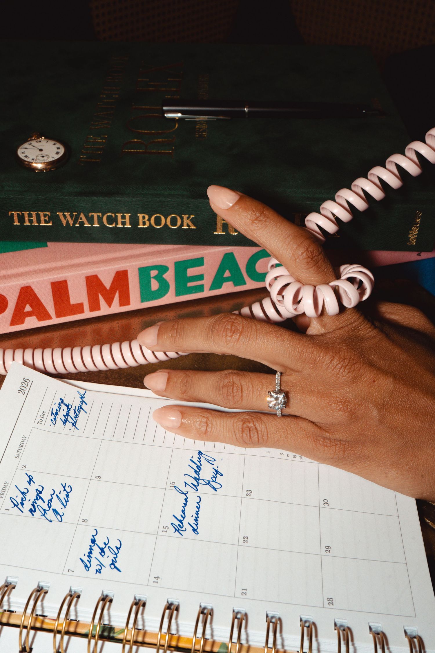 A diamond ring on a hand resting on a calendar beside Palm Beach and watch books.