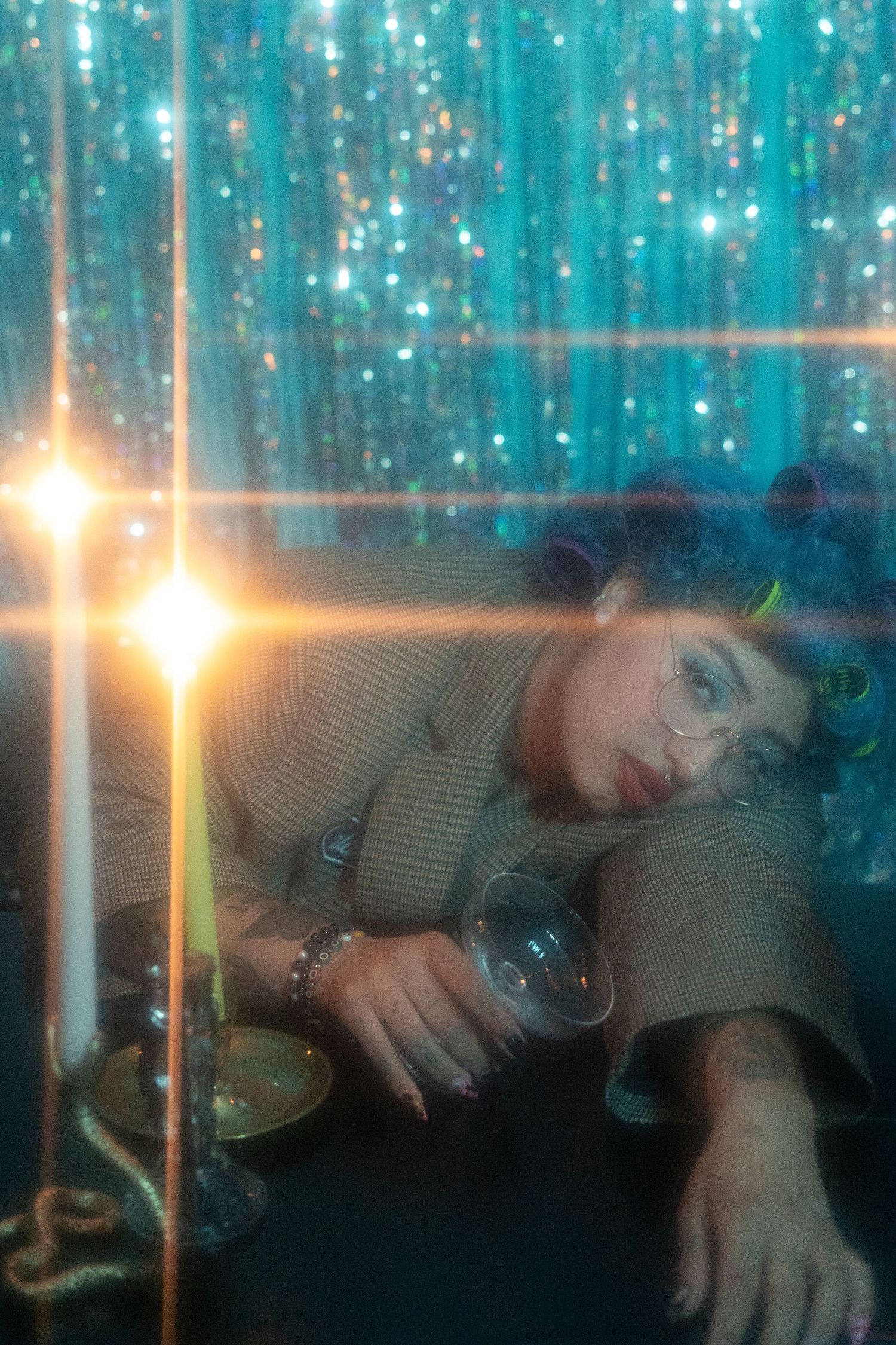 Tattooed woman with blue curly hair and glasses reclines on a table by candlelight against a teal glitter curtain backdrop.