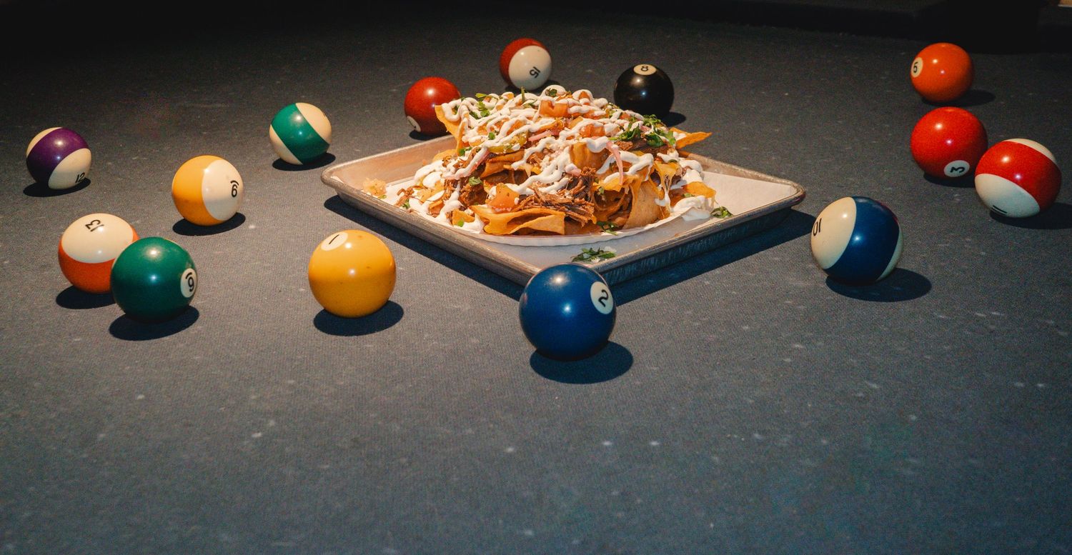 Loaded nachos on a square plate surrounded by scattered colorful billiard balls on a pool table.