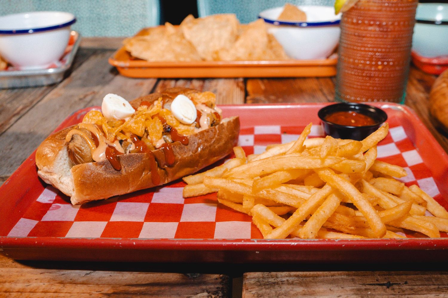 A loaded hot dog topped with caramelized onions served with golden crispy french fries on a red checkered tray.