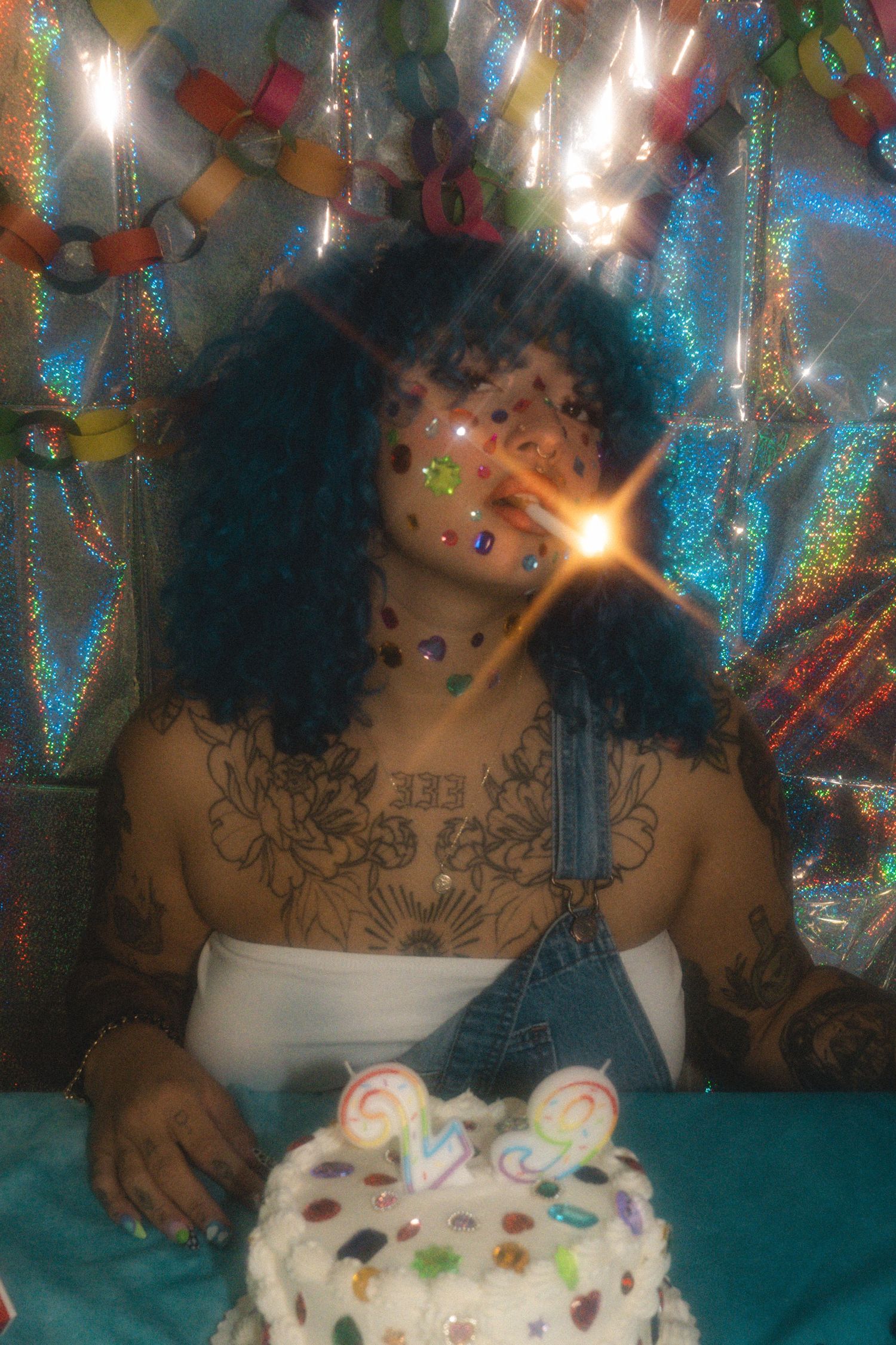 Person with curly hair, face gems, and chest tattoos sits before a decorated birthday cake with candles.