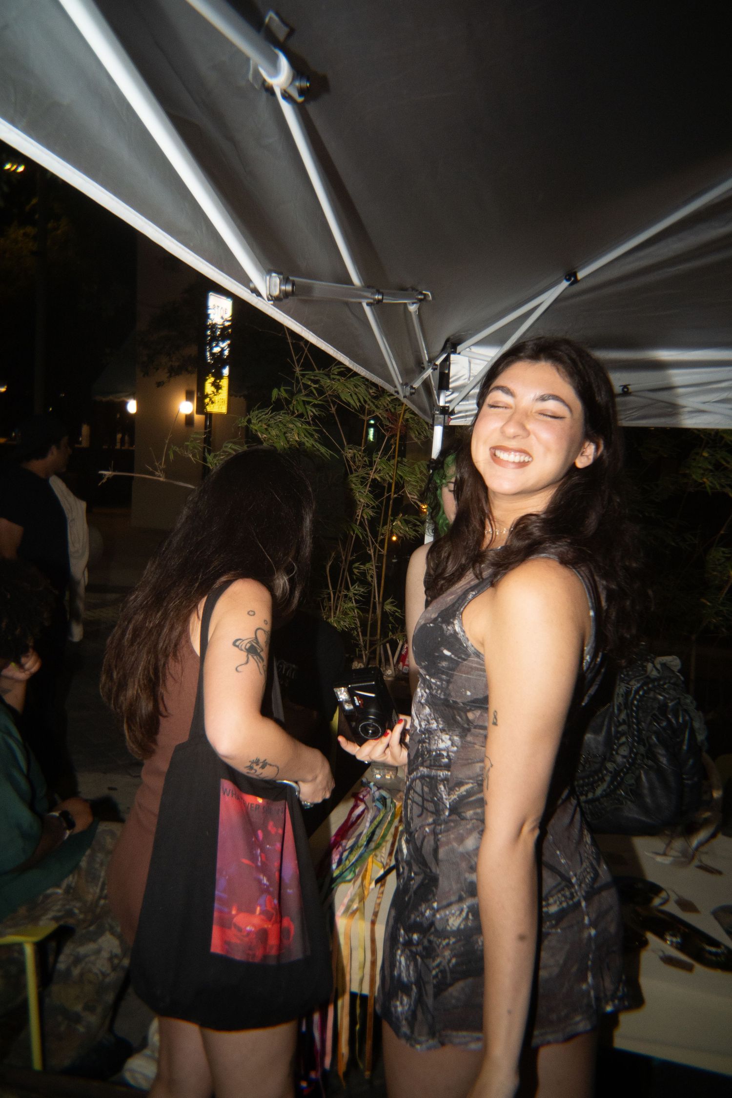 Two young women smiling and socializing at an outdoor evening event under a white canopy tent.