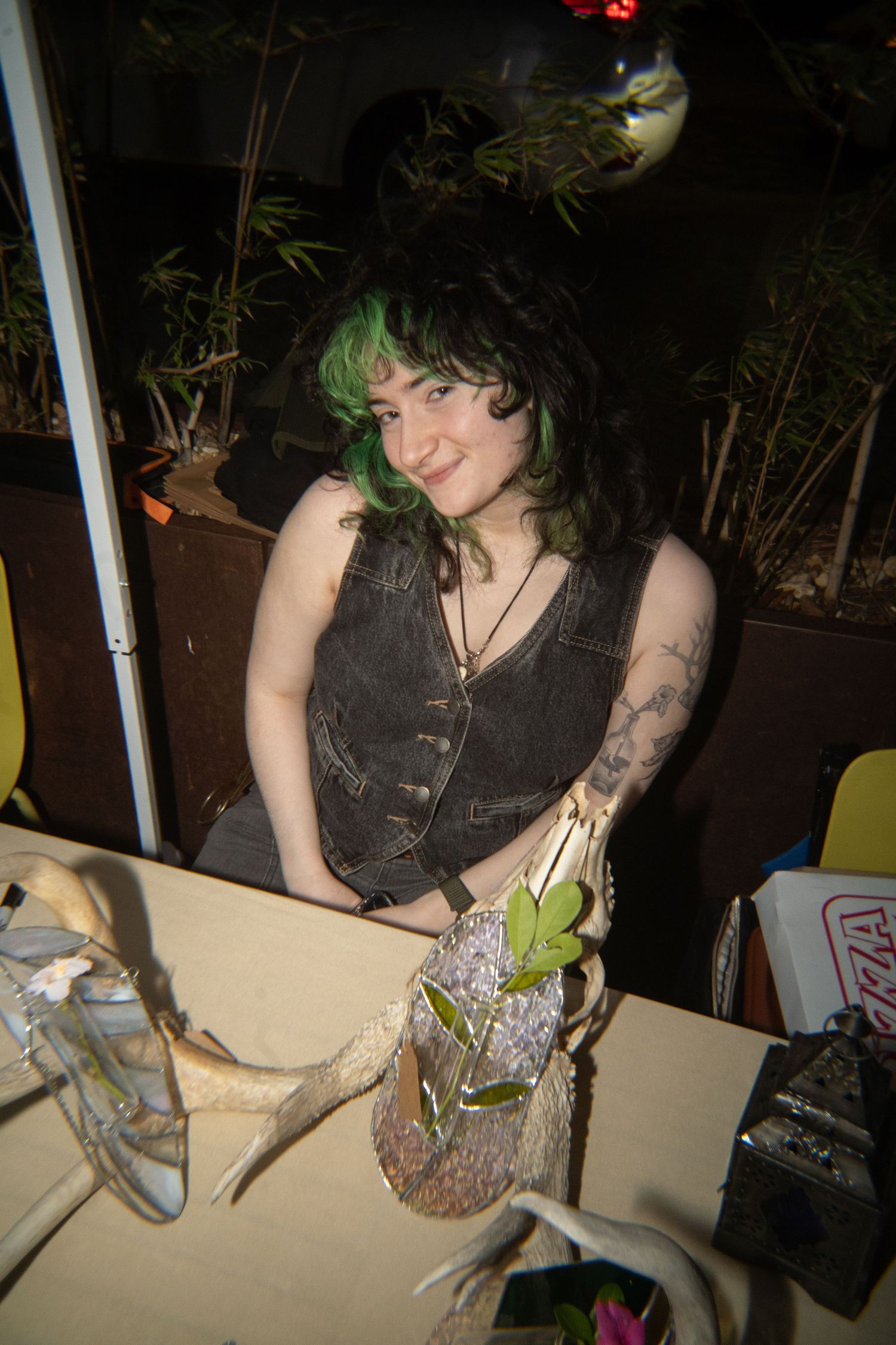 Young woman with green-tipped black hair poses with animal skull and antlers at a decorated table indoors.
