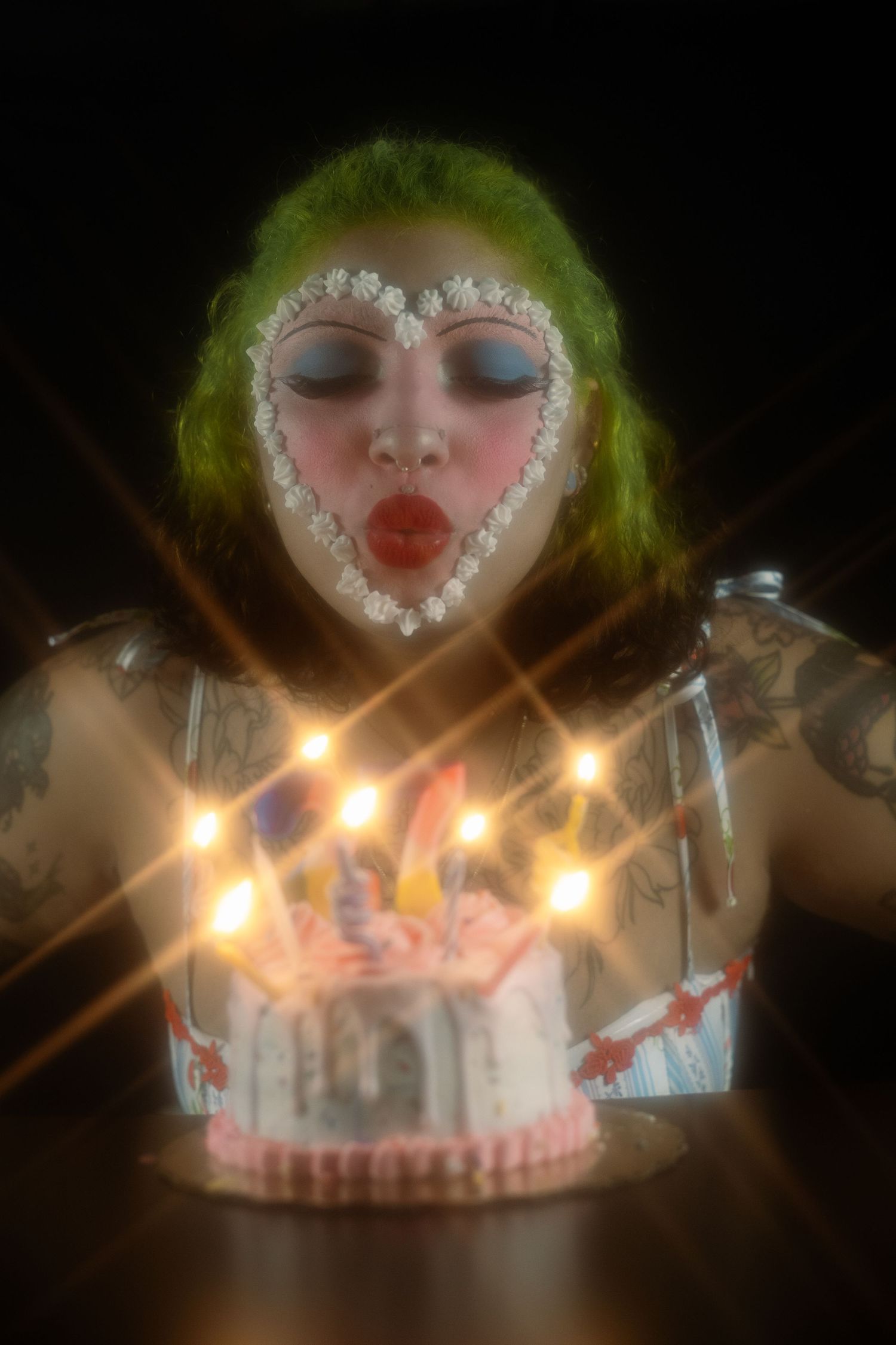Tattooed woman with green hair and heart face paint blowing out glowing candles on a birthday cake.
