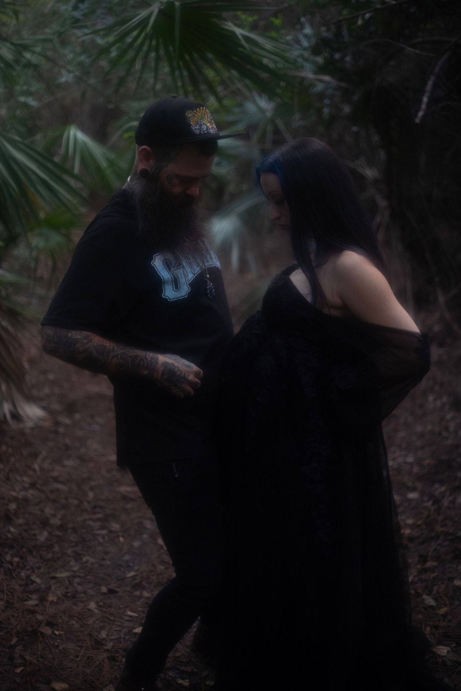 Couple sharing a tender moment in a dark forest, man with beard and tattoos, woman in black gown.