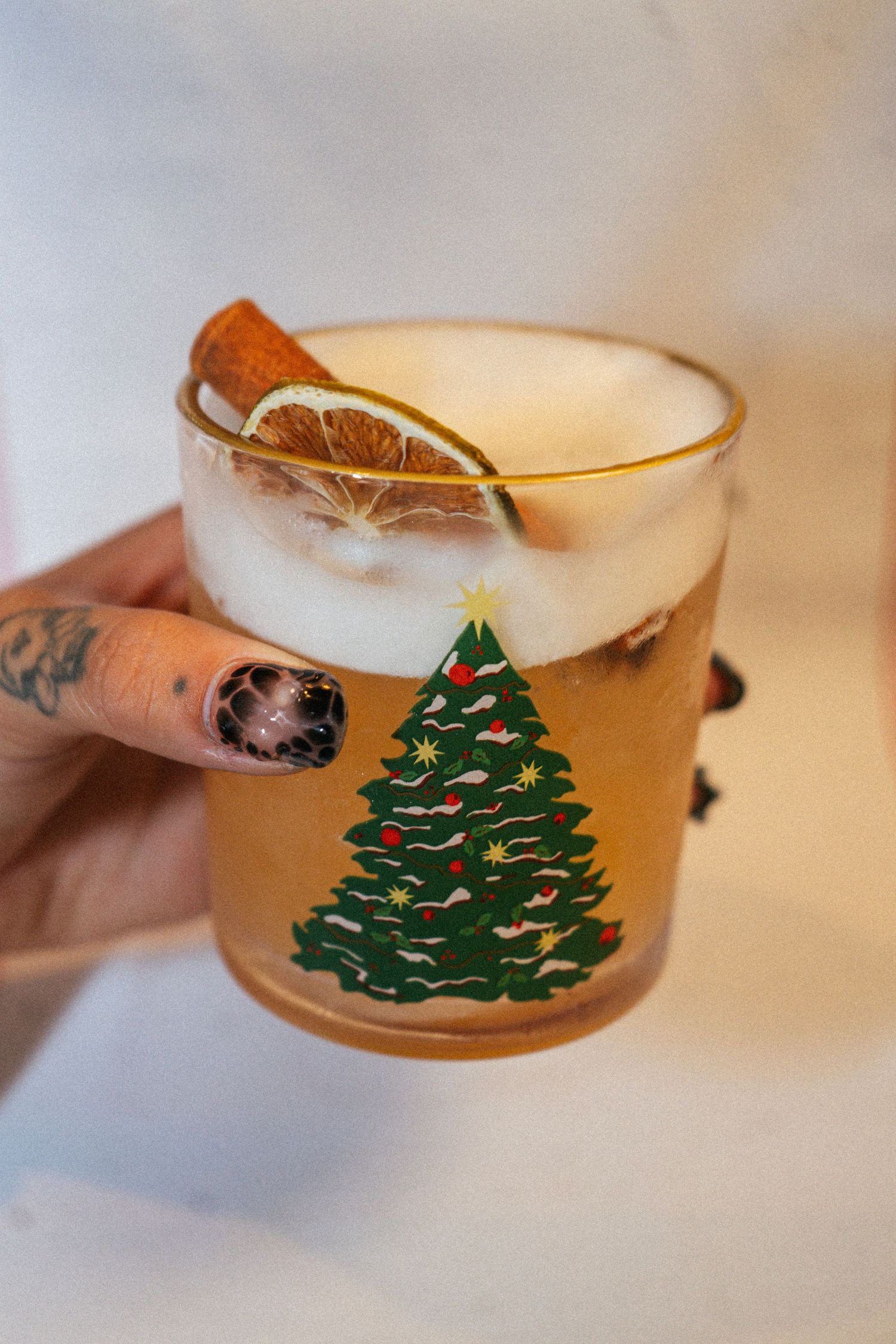 Hand with festive nail art holding a Christmas tree rocks glass cocktail garnished with orange and cinnamon.