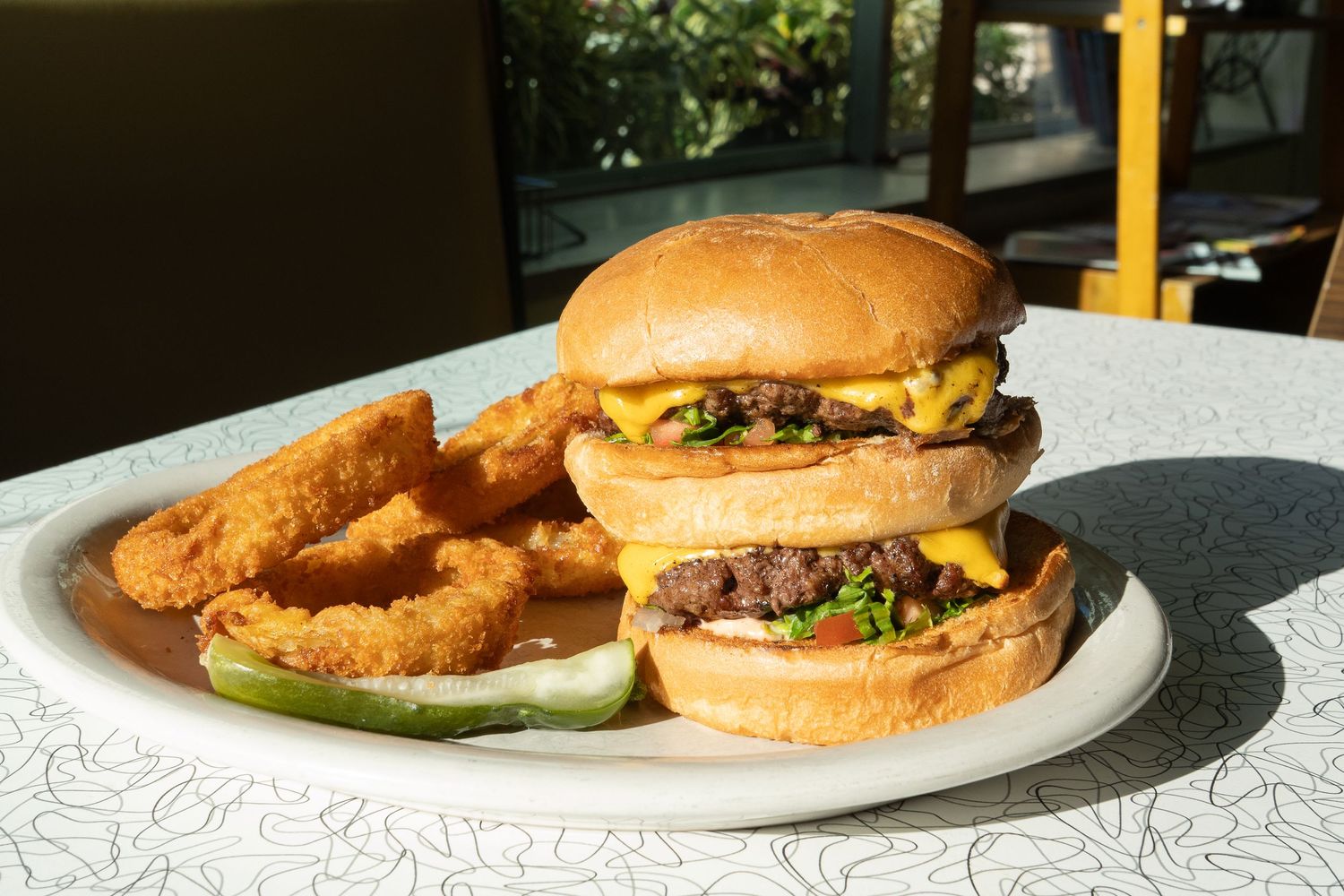 Double cheeseburger with melted cheese, fresh toppings, crispy onion rings, and pickles served on a white plate.