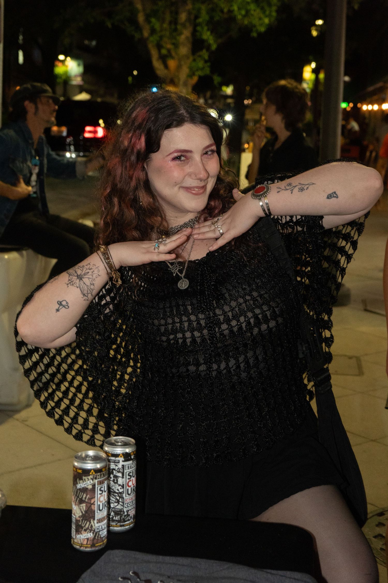 Woman with tattoos in black crochet top smiling at outdoor night event with drinks on table.