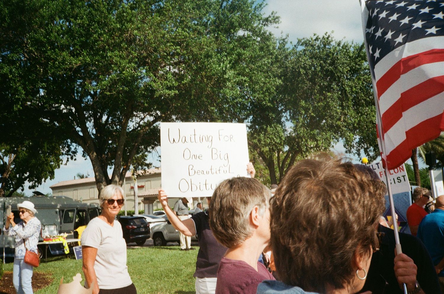 Protesters gather on grass holding signs and American flag at outdoor political demonstration.