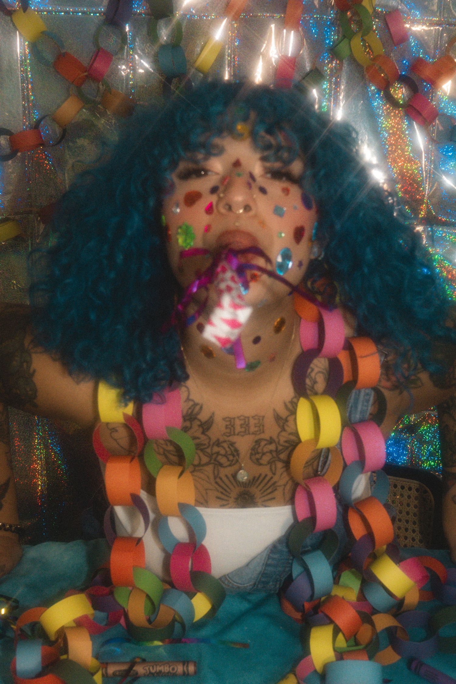 Person with teal afro wig, confetti, tattoos, and colorful paper chain garlands in a festive, glittery setting.