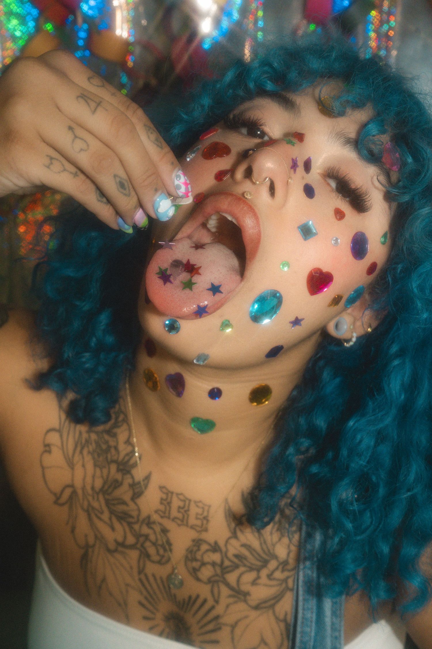 Woman with teal curly hair, face gems, tattoos, and tongue out posing at a colorful glitter party.