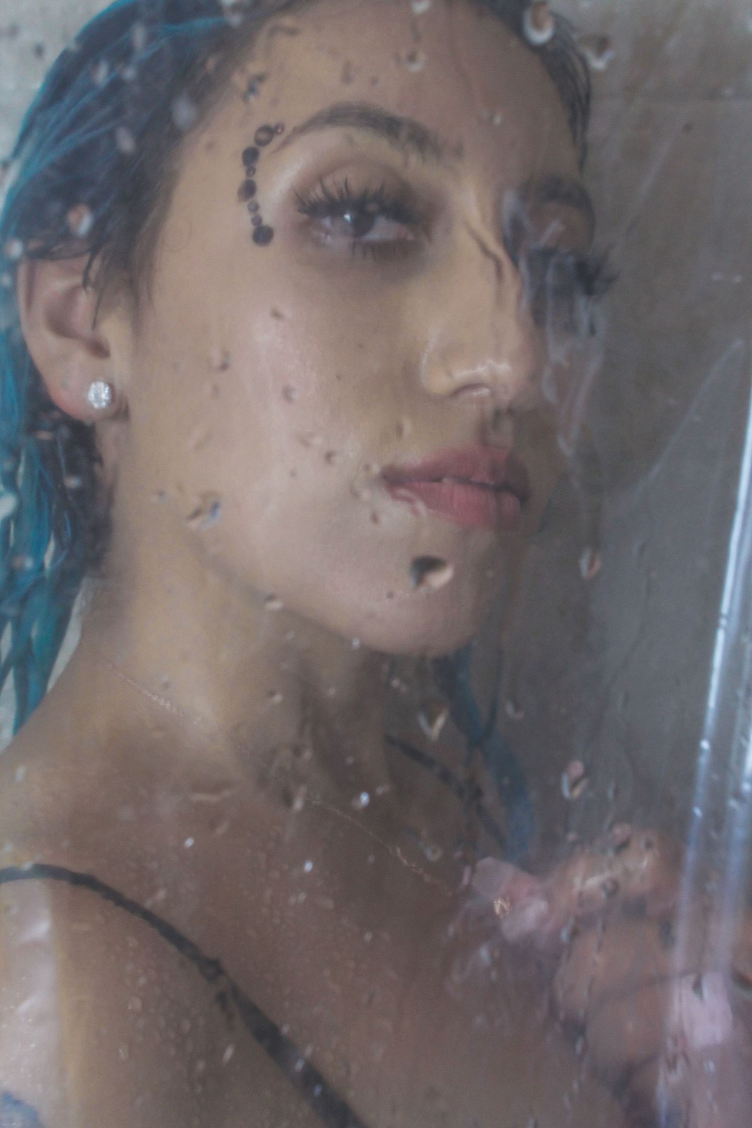 A woman with blue hair and black face gems presses against a wet glass shower door, water droplets scattered across.