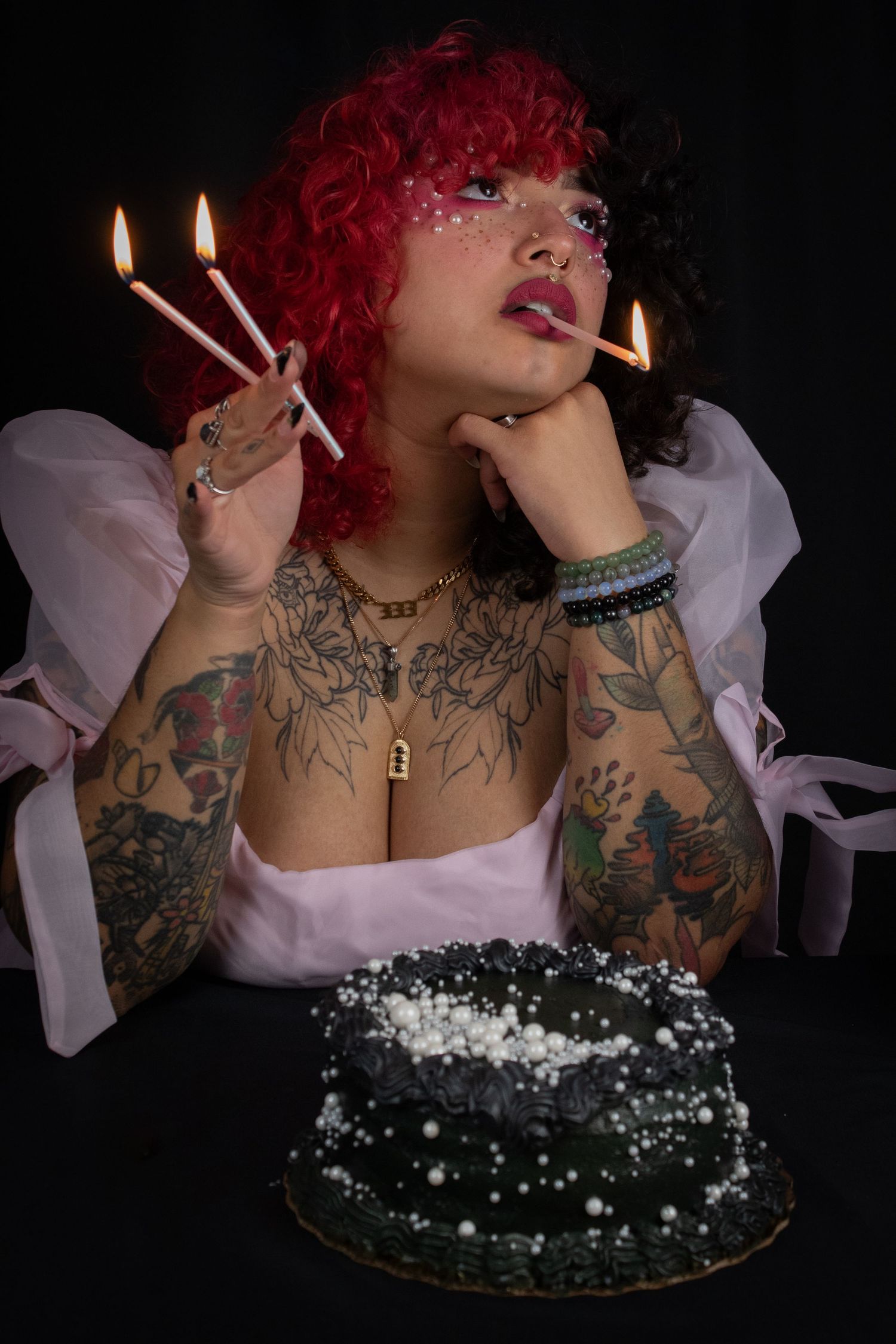 Tattooed woman with red and black curly hair poses with birthday candles and dark cake.