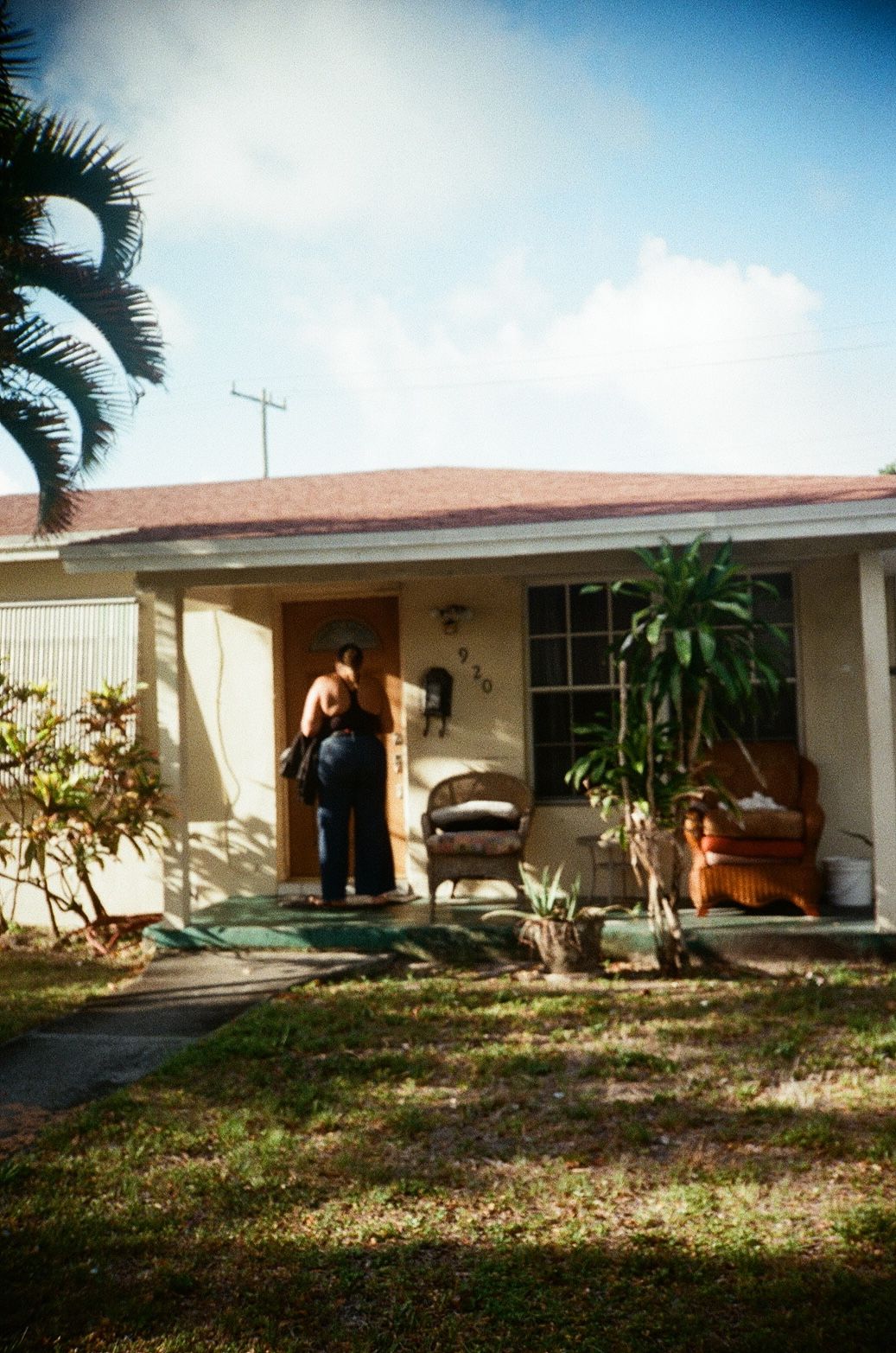 Person standing at the front door of a single-story Florida home with wicker chairs and tropical plants.