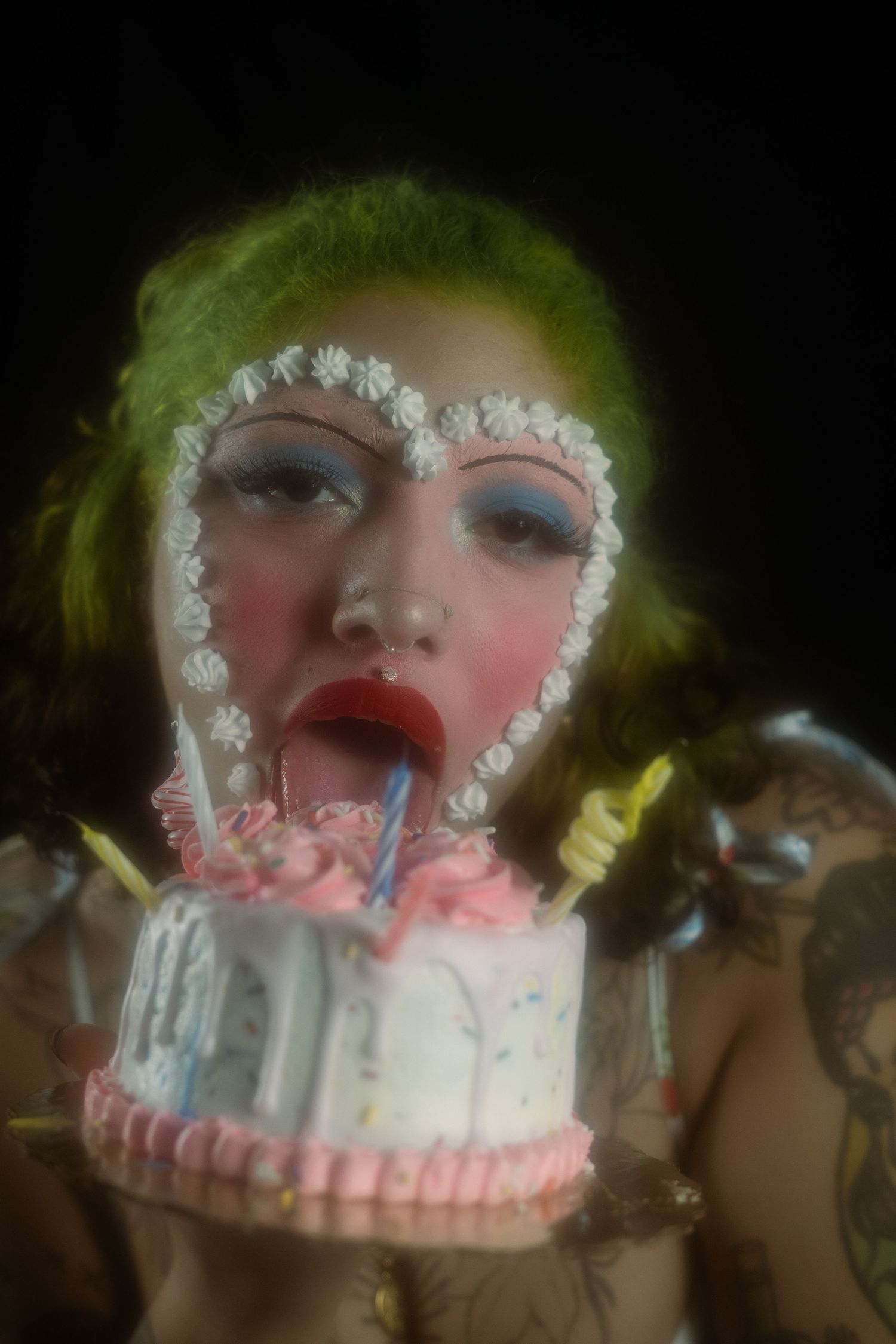 Tattooed woman with green hair and heart face paint licking a decorated birthday cake with candles.
