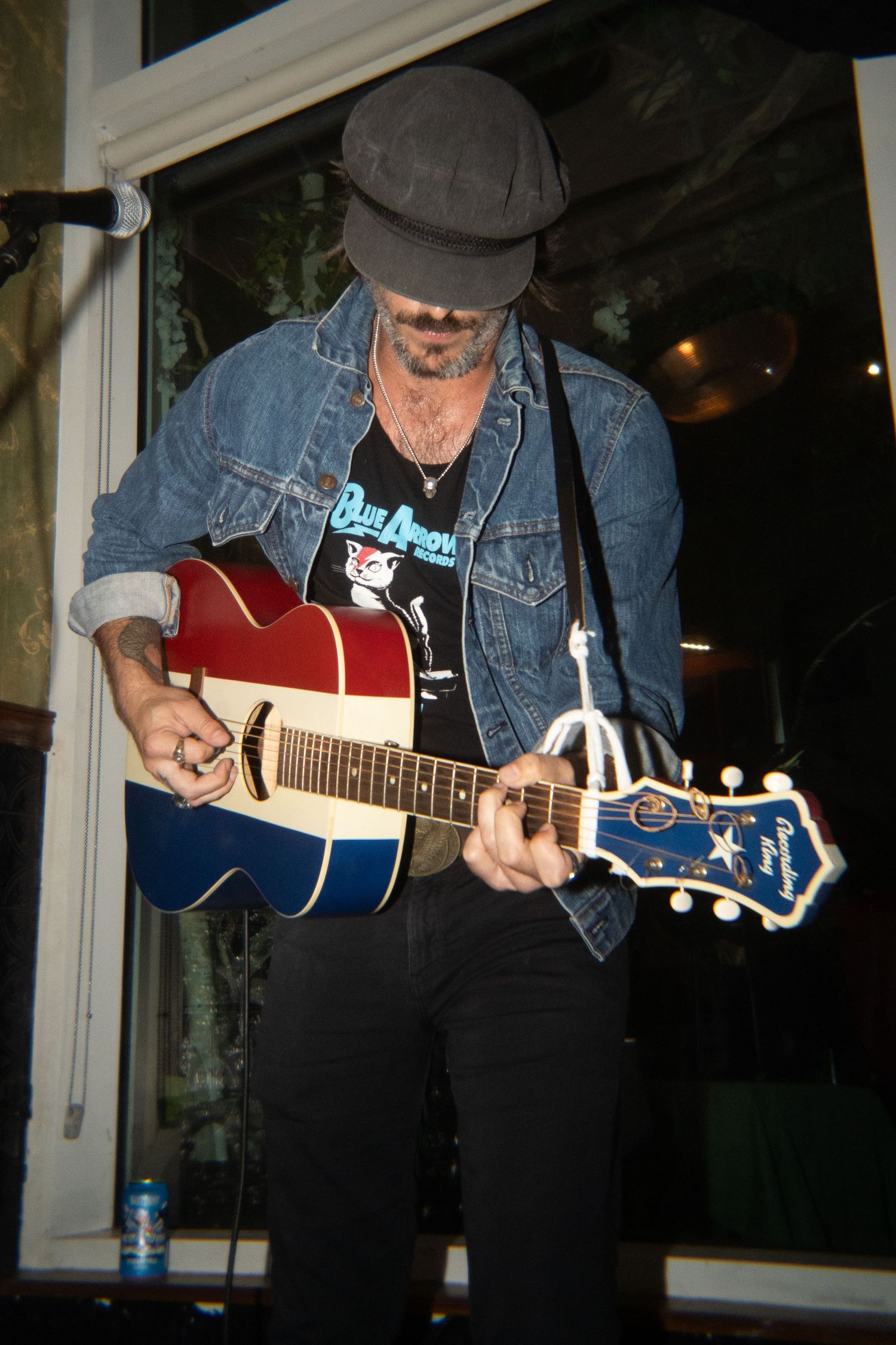 Man in denim jacket and flat cap playing a red, white, and blue acoustic guitar on stage.