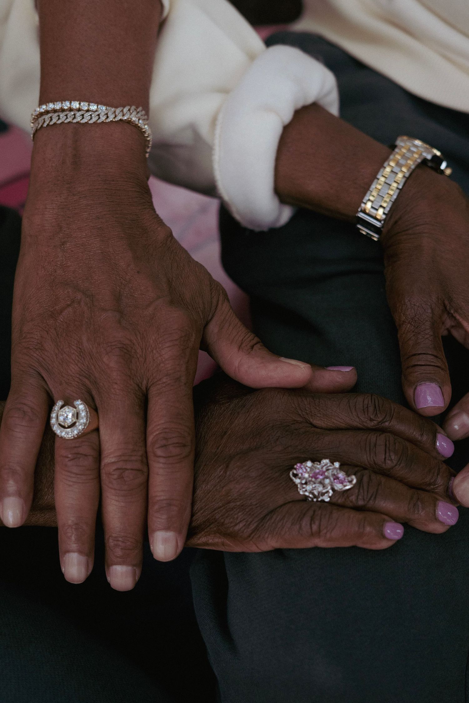 Two elderly Black women's hands adorned with diamond rings and bracelets resting together.