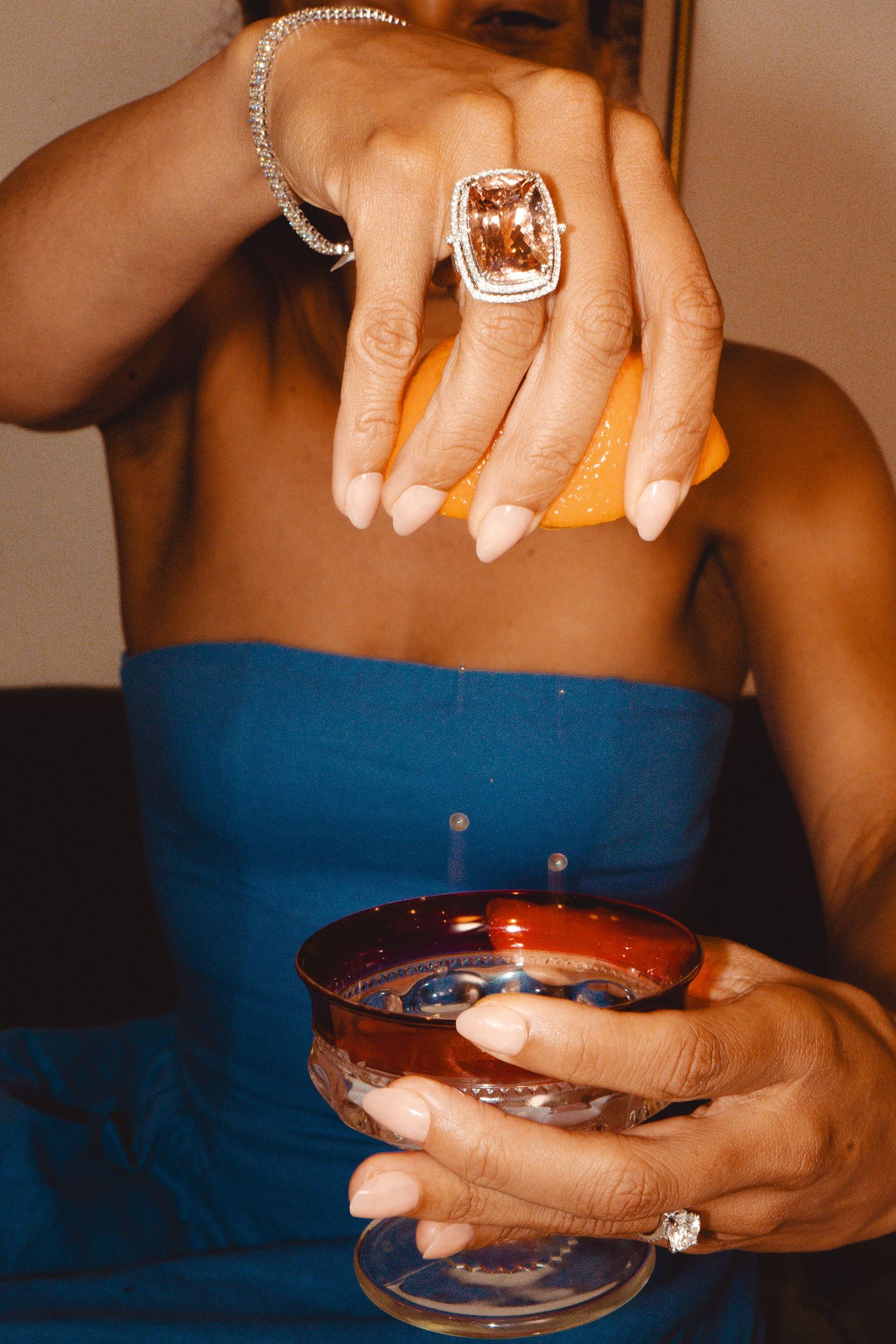 Woman in blue dress squeezing lemon into a cocktail glass while wearing a large statement ring.