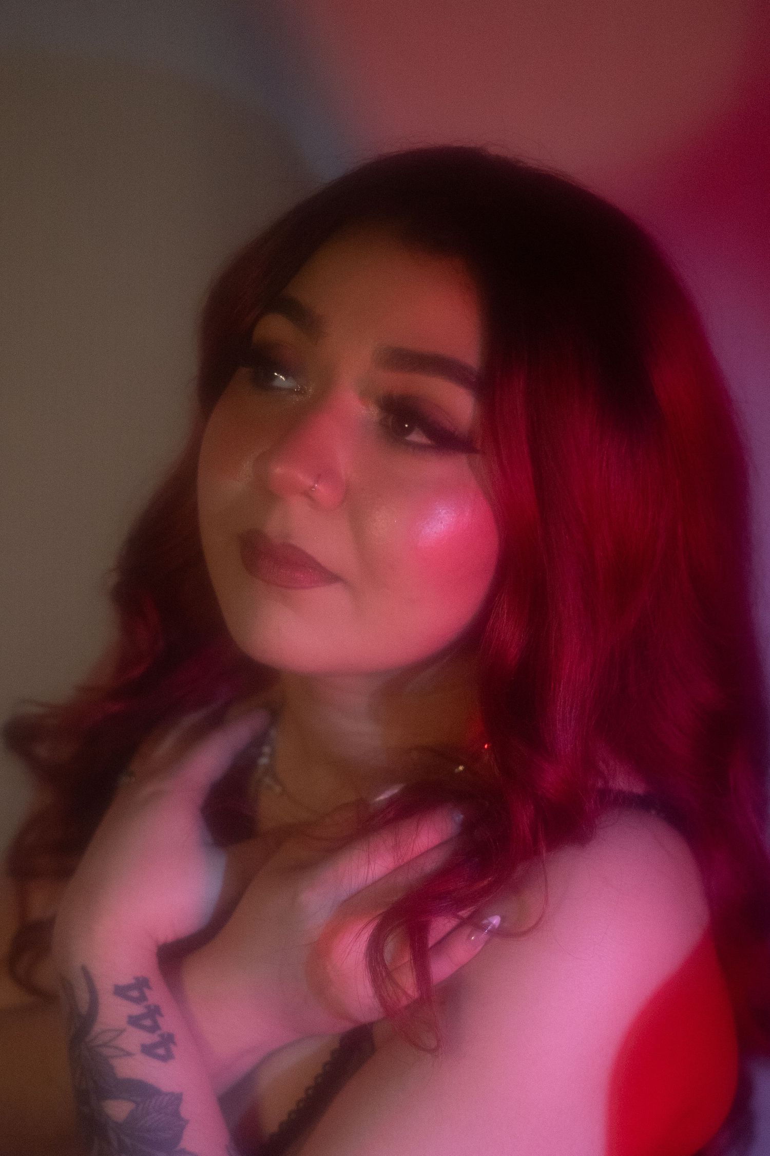 Woman with red wavy hair and arm tattoo posing in soft pink and red lighting.