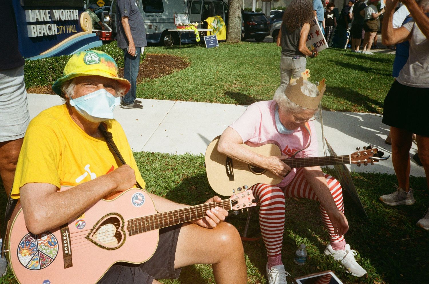 Two musicians wearing masks play acoustic guitars outdoors at a community gathering on a sunny day.