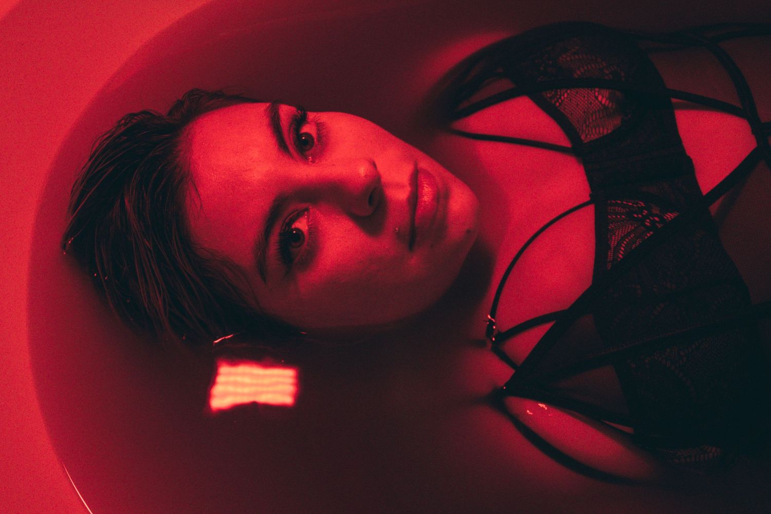 A woman in black lace lingerie lies submerged in a bathtub illuminated by dramatic red lighting.