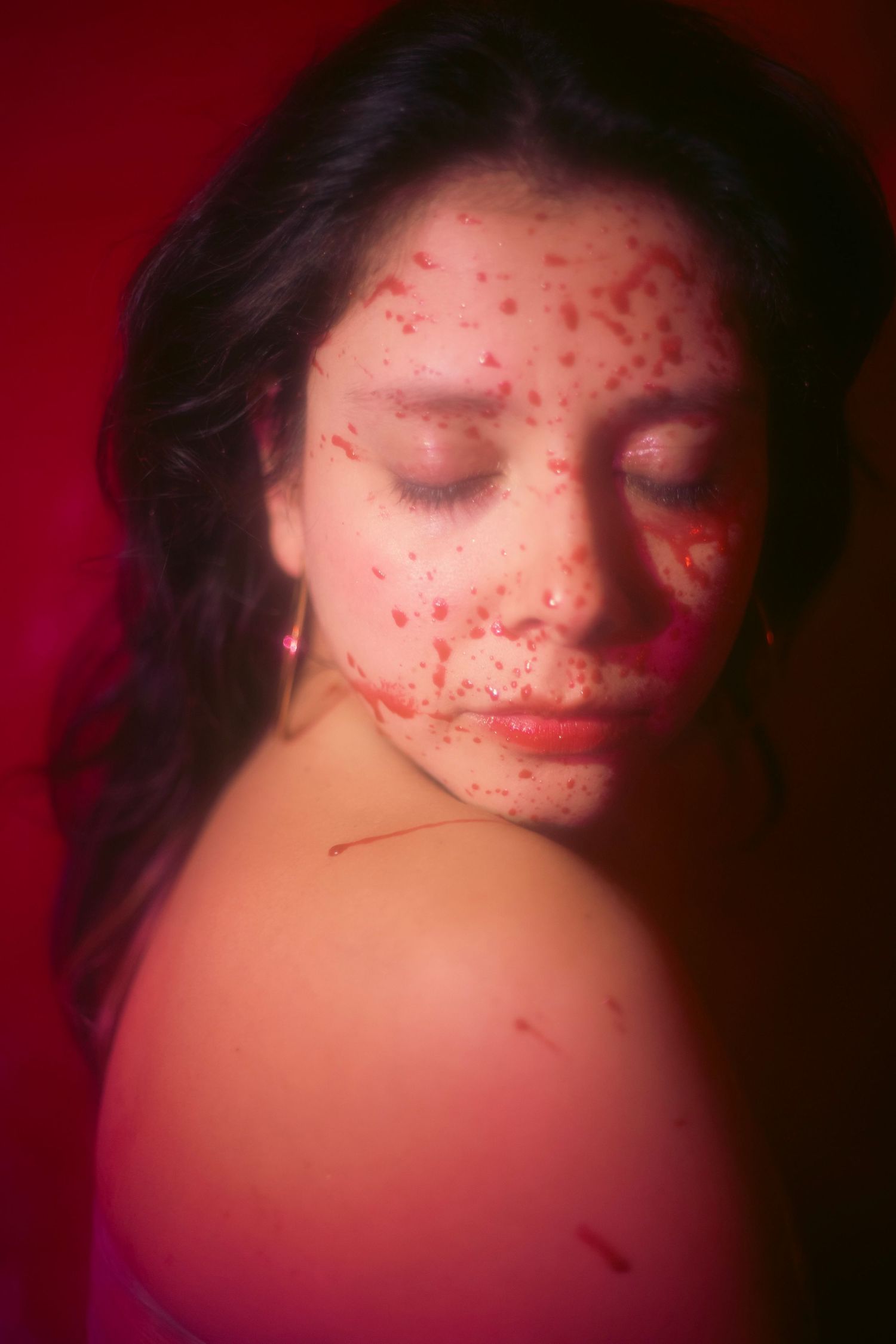 A woman with dark hair and simulated blood splatter on her face poses in a red-lit horror shoot.