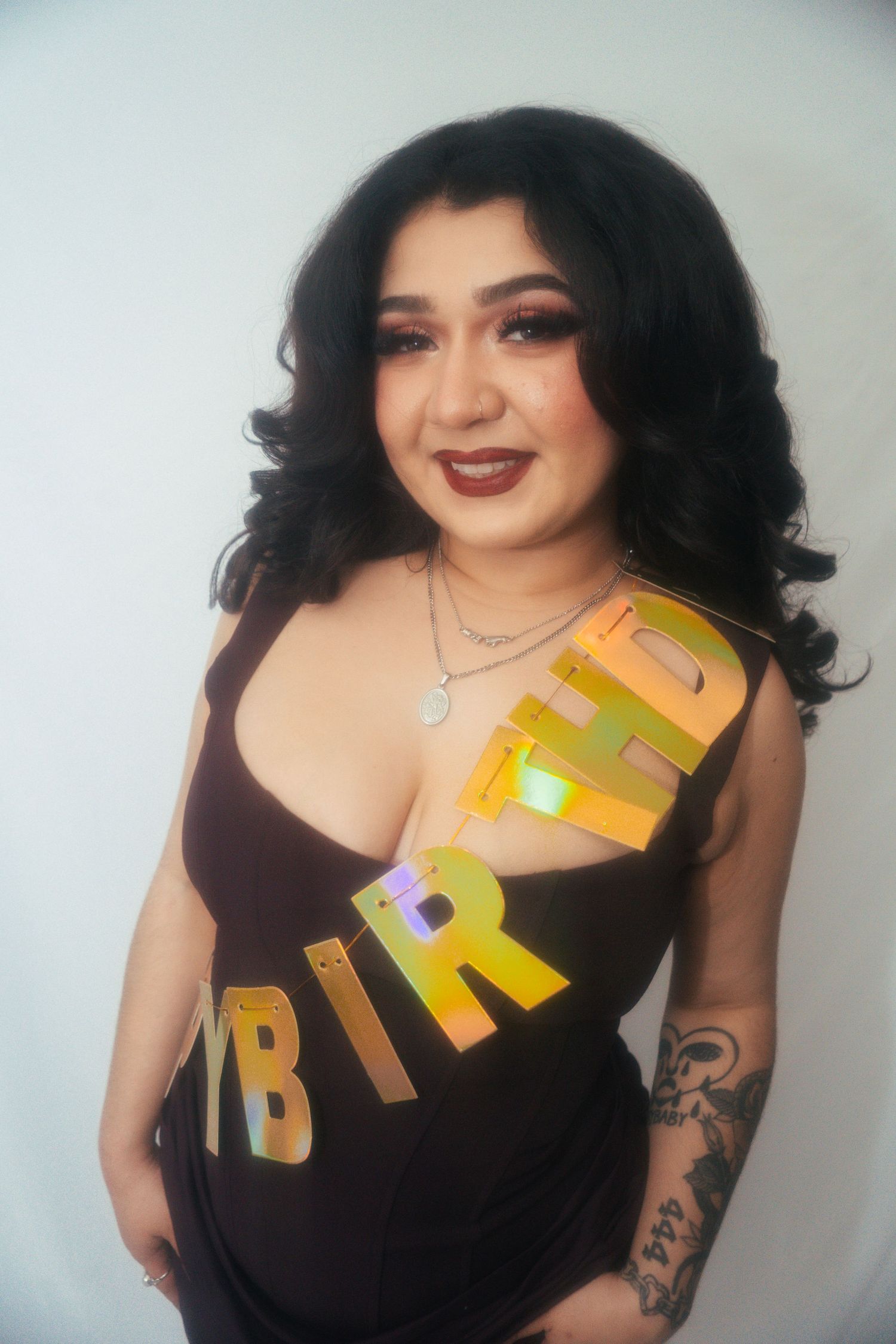 Woman in black dress wearing gold Happy Birthday sash smiling against white background.