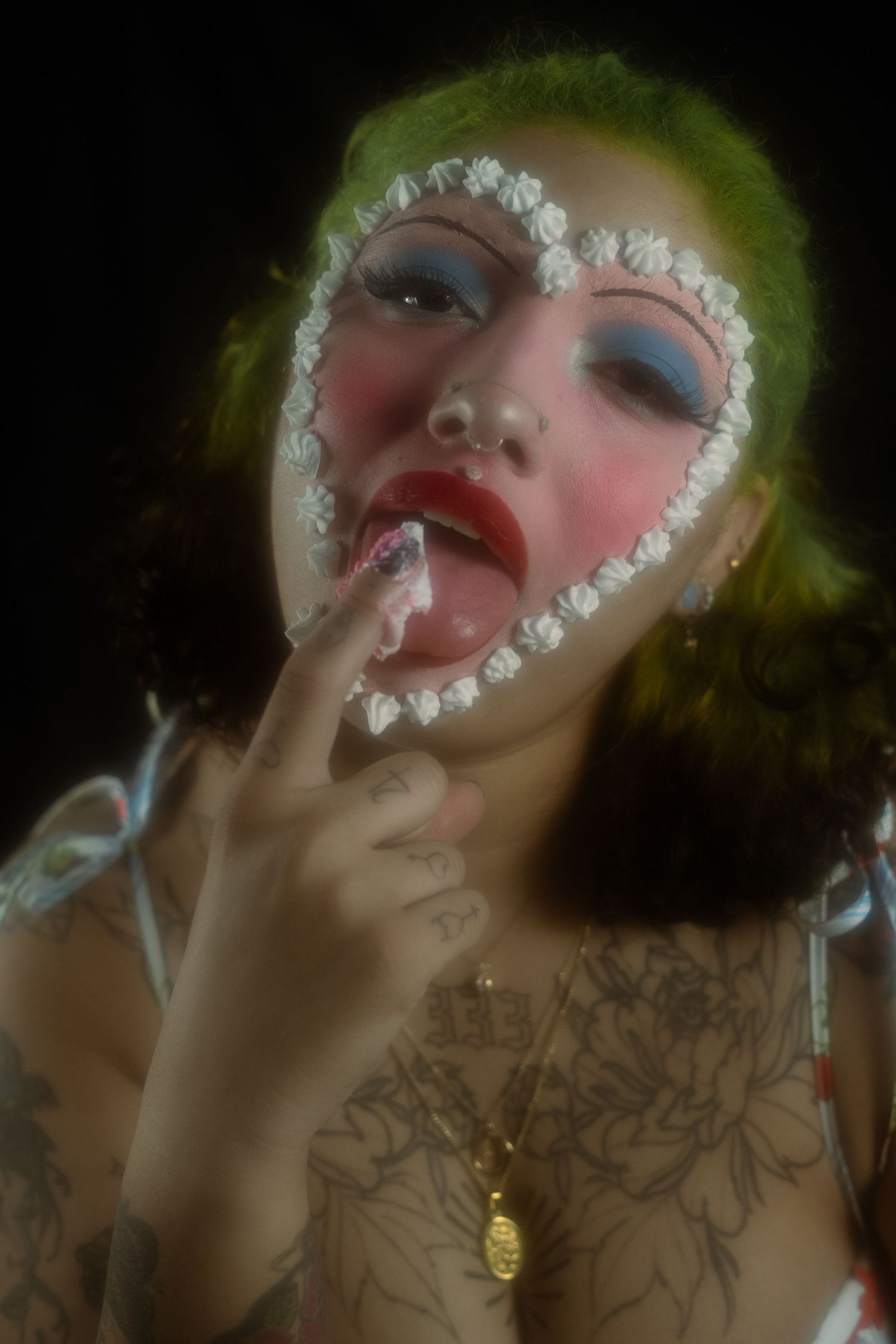 Tattooed woman with green hair and heart-shaped lace face paint licking frosting from her finger.