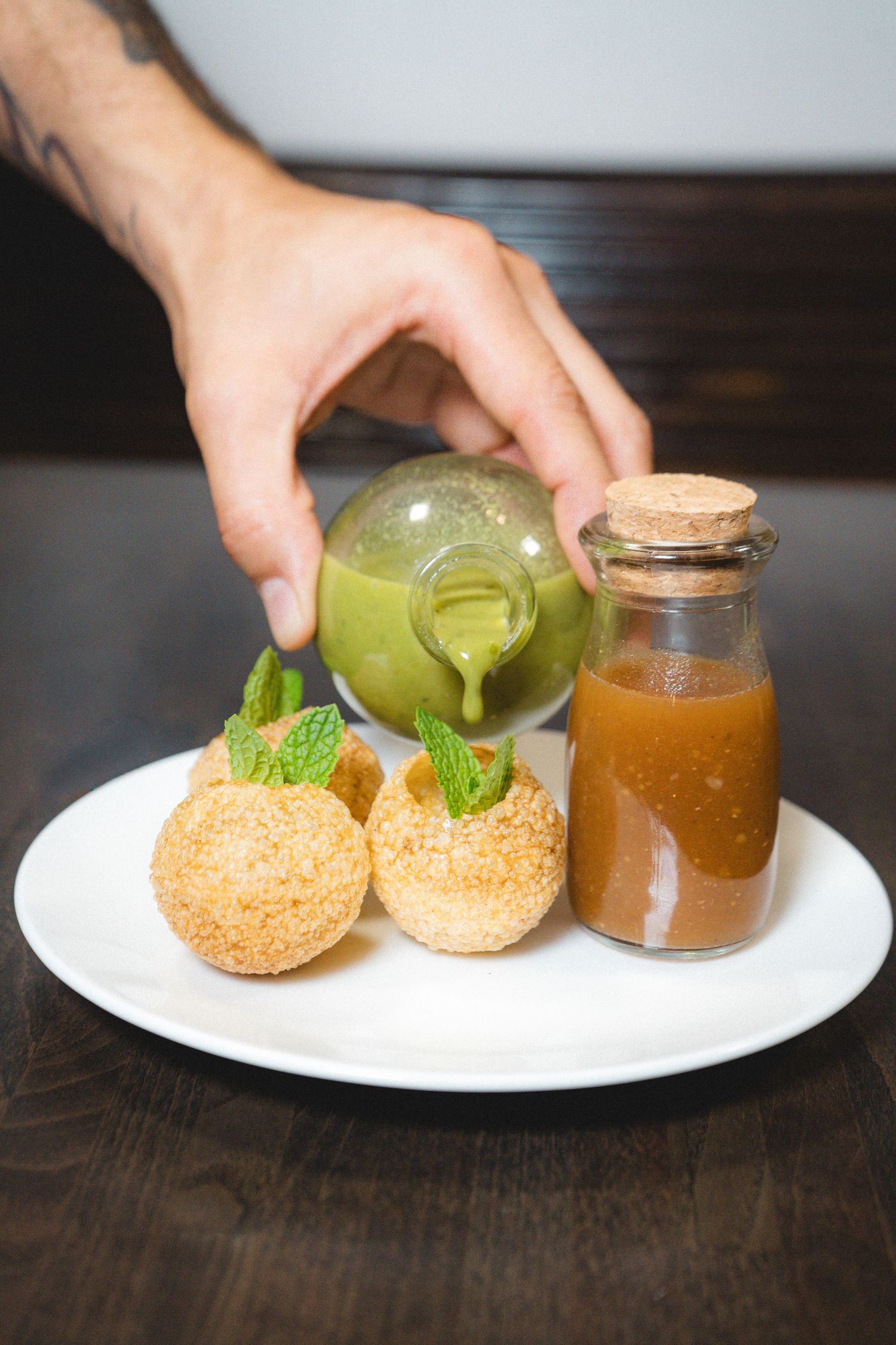 Hand pouring green sauce over crispy fried balls garnished with mint on white plate with caramel sauce bottle.