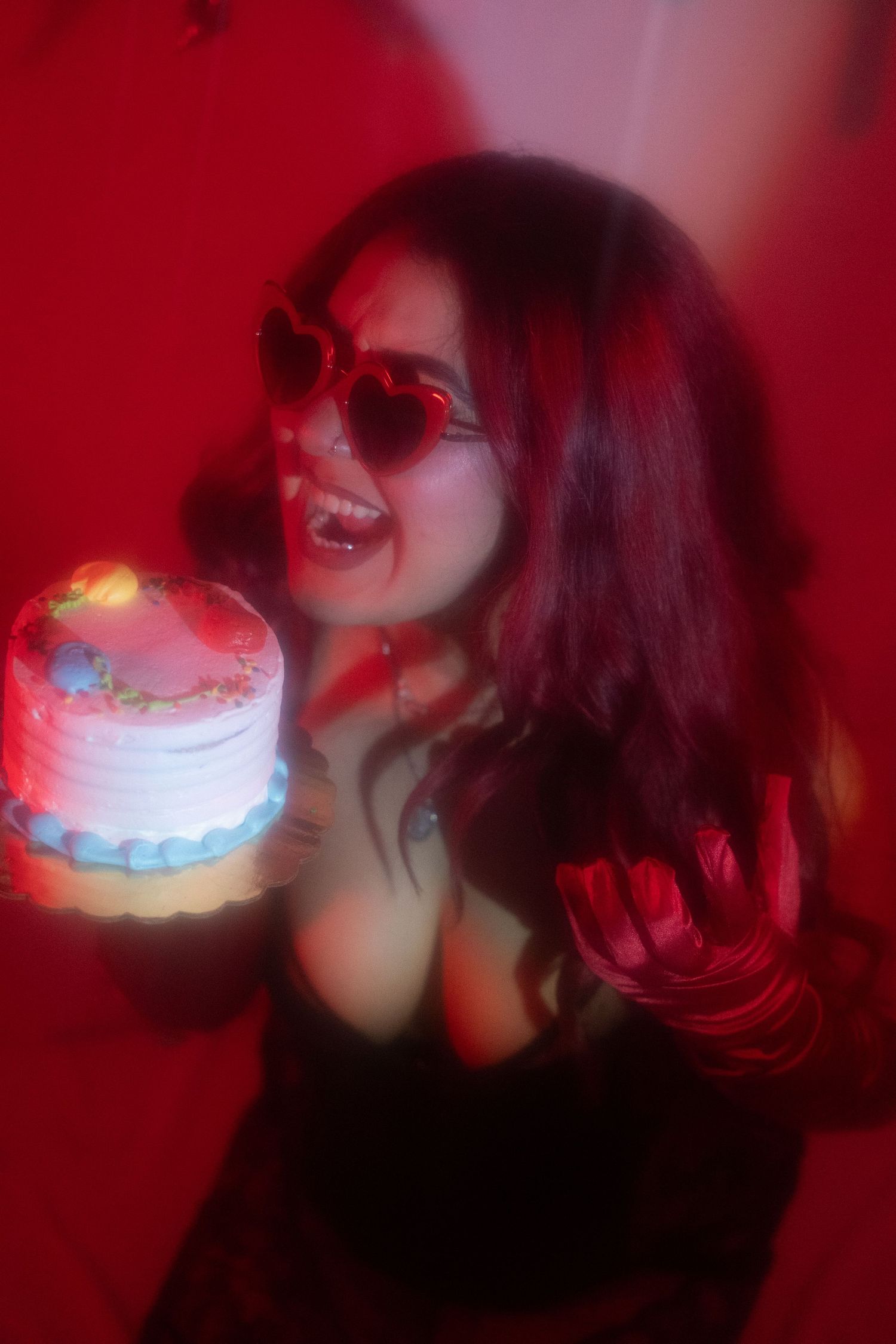 Woman with red hair wearing heart sunglasses holding a glowing birthday cake in red-lit setting.