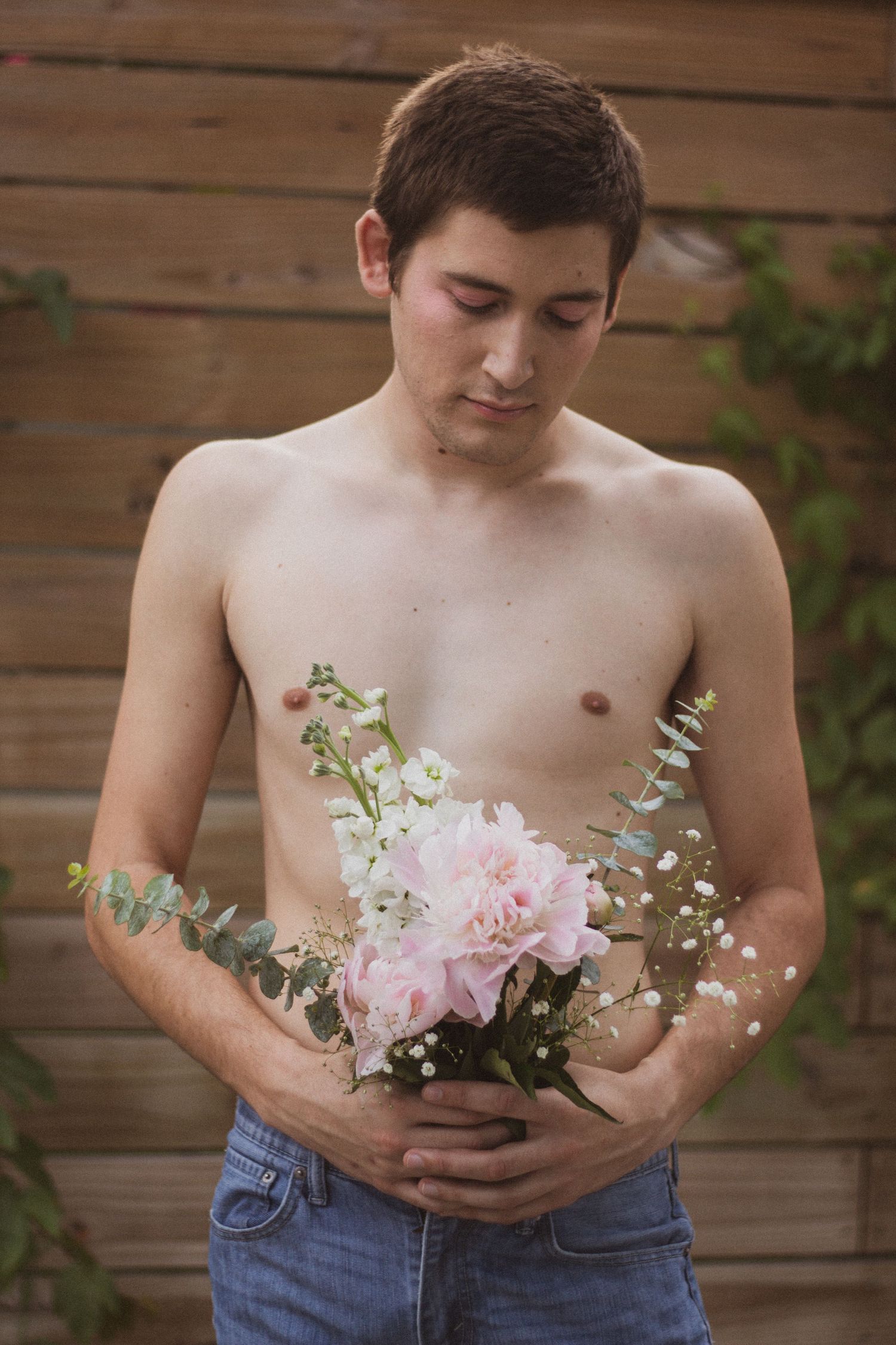 A shirtless young man gazes down while holding a delicate bouquet of pink peonies and white flowers outdoors.