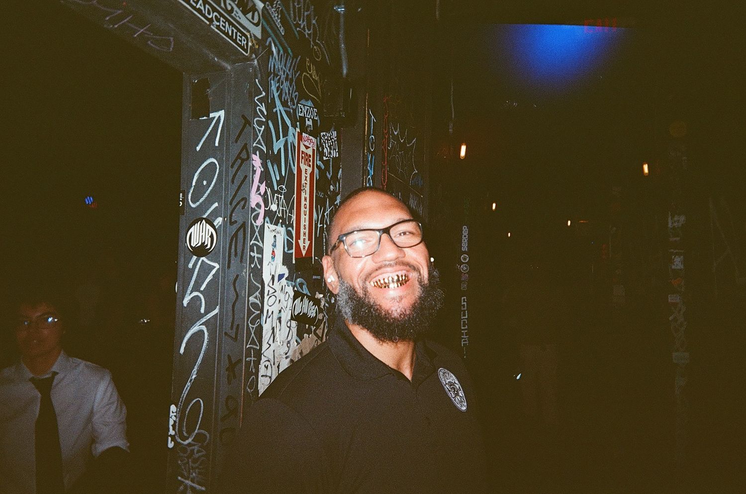 A smiling man with glasses and a beard laughs joyfully at night near a graffiti-covered wall.
