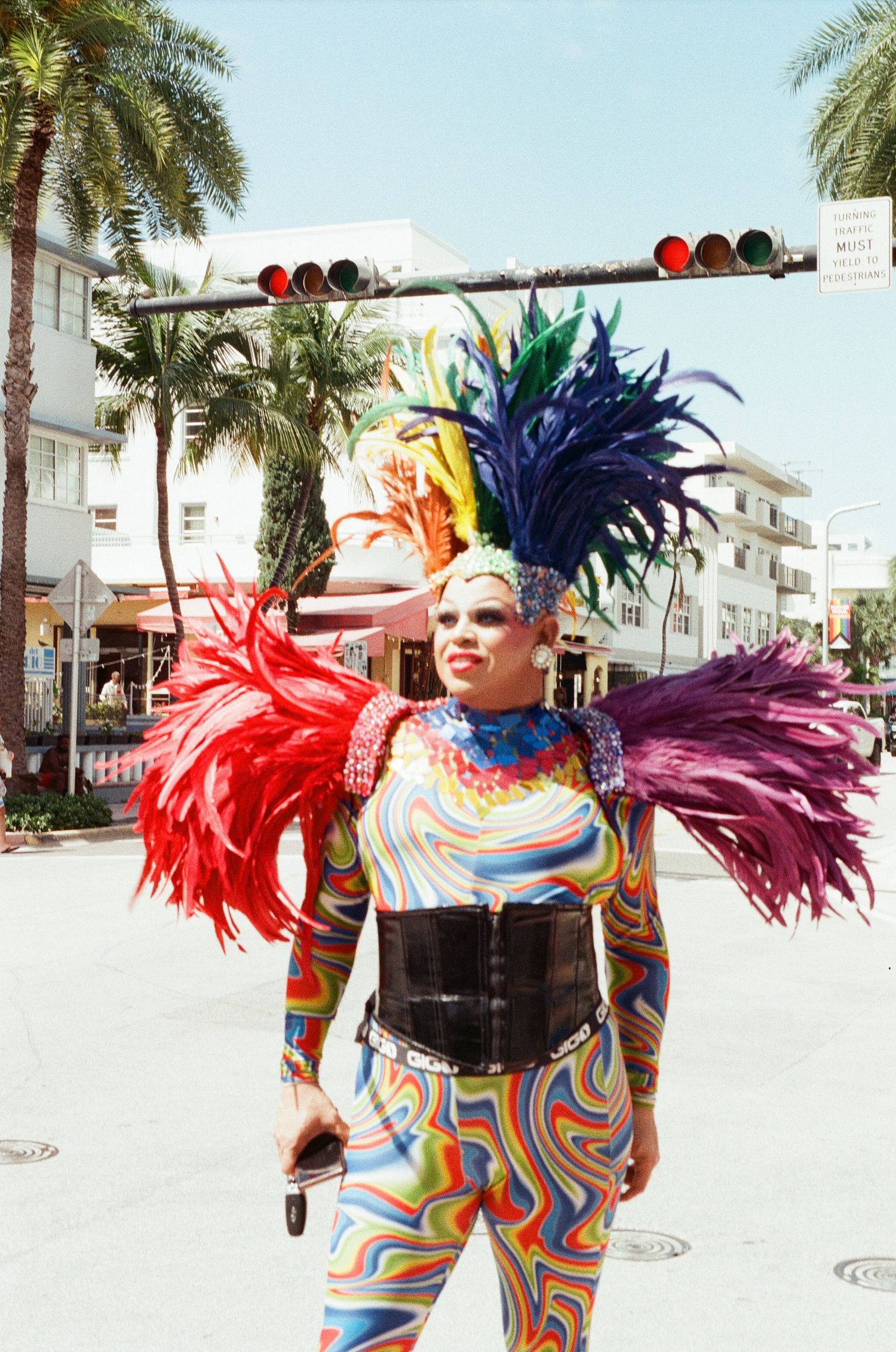 Colorful drag performer in rainbow bodysuit with feathered headdress and shoulder pieces poses on a sunny Miami street.