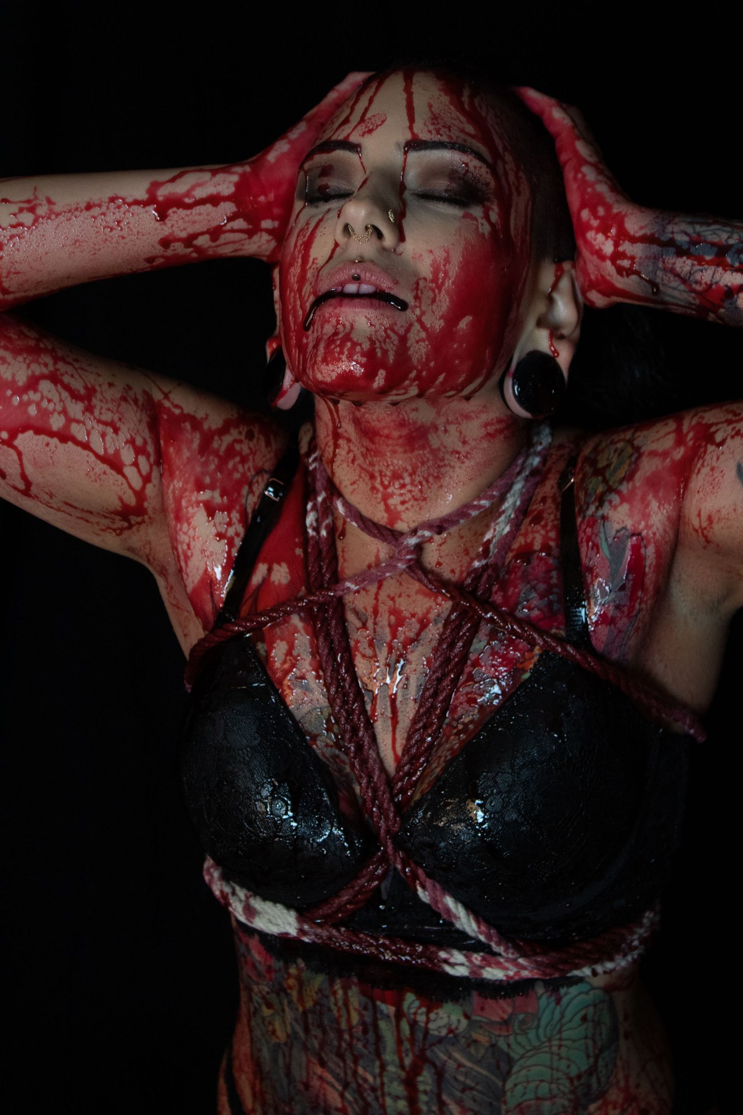 Woman with tattoos covered in red paint simulating blood, wearing black bra with rope harness in dark horror art photography.