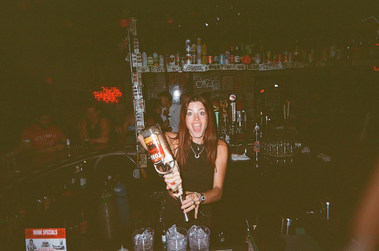 Female bartender smiling while pouring a drink at a dimly lit bar with bottles on shelves in the background.