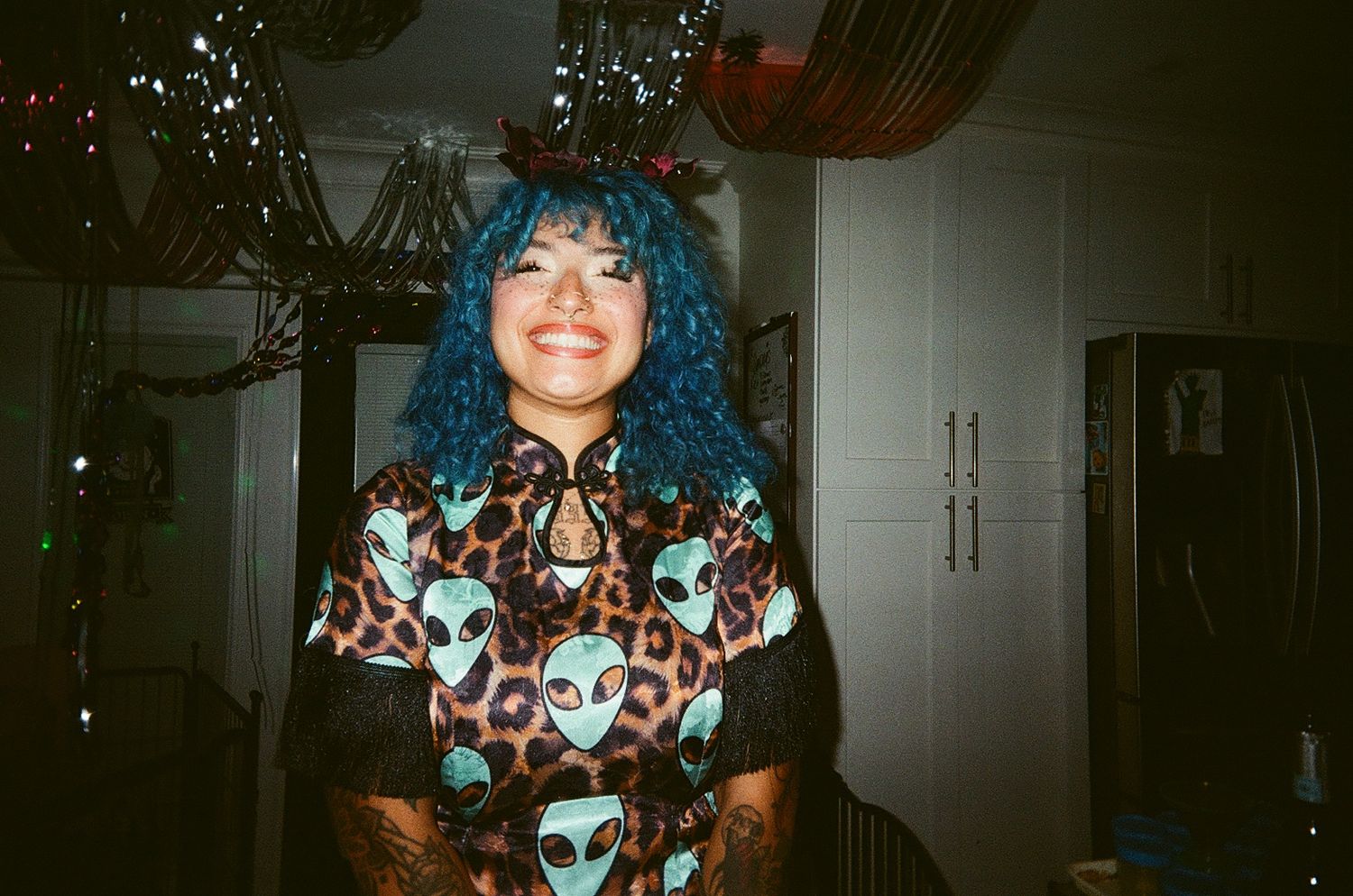Woman with blue curly hair smiling, wearing alien print leopard top, festive holiday decorations in background.