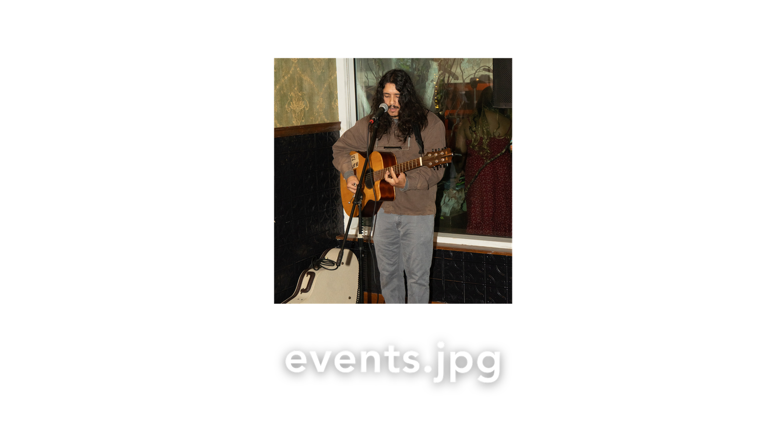 A musician with long hair plays acoustic guitar while standing indoors at a live music event.