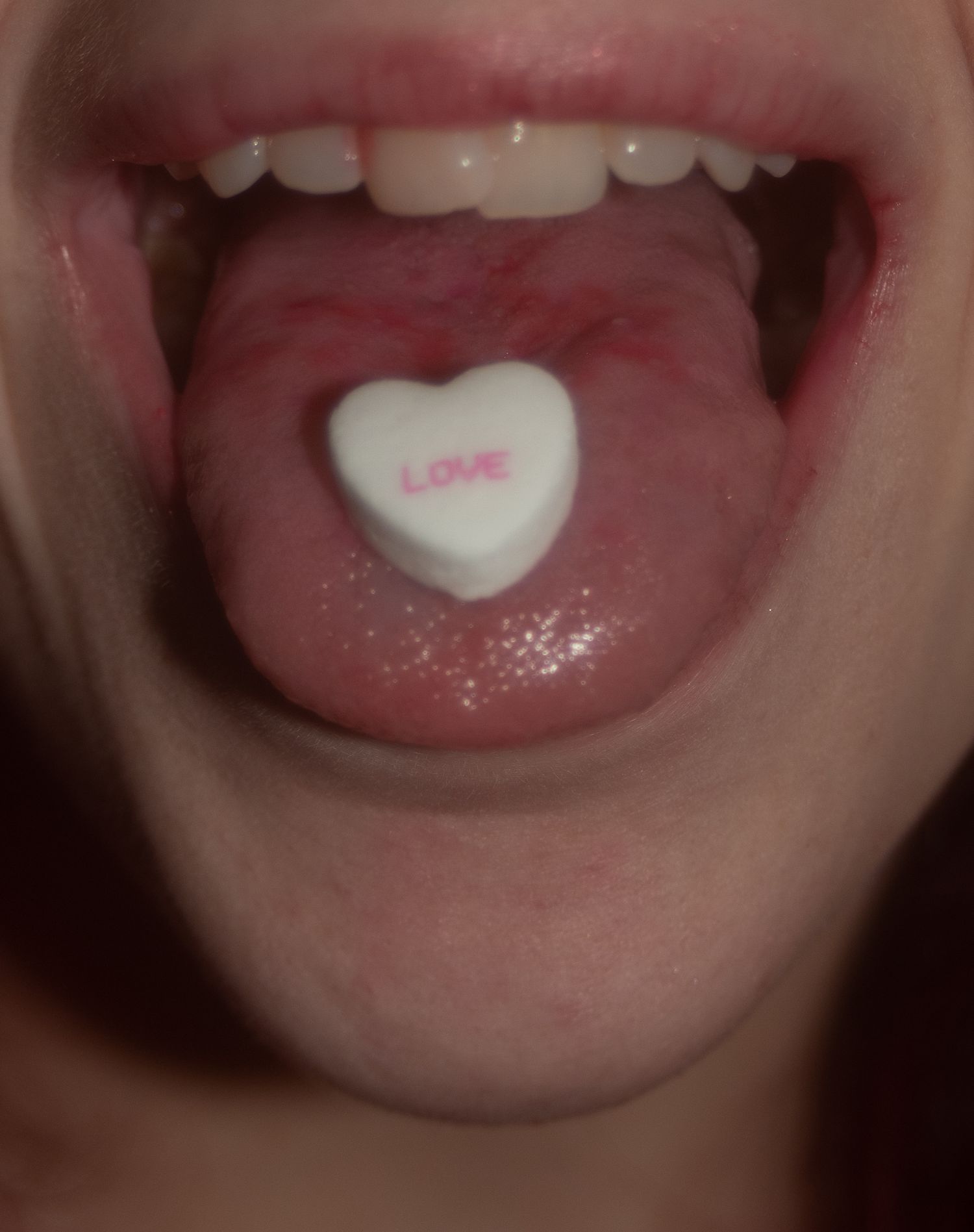 Close-up of open mouth with a white heart-shaped candy printed with LOVE resting on the tongue.