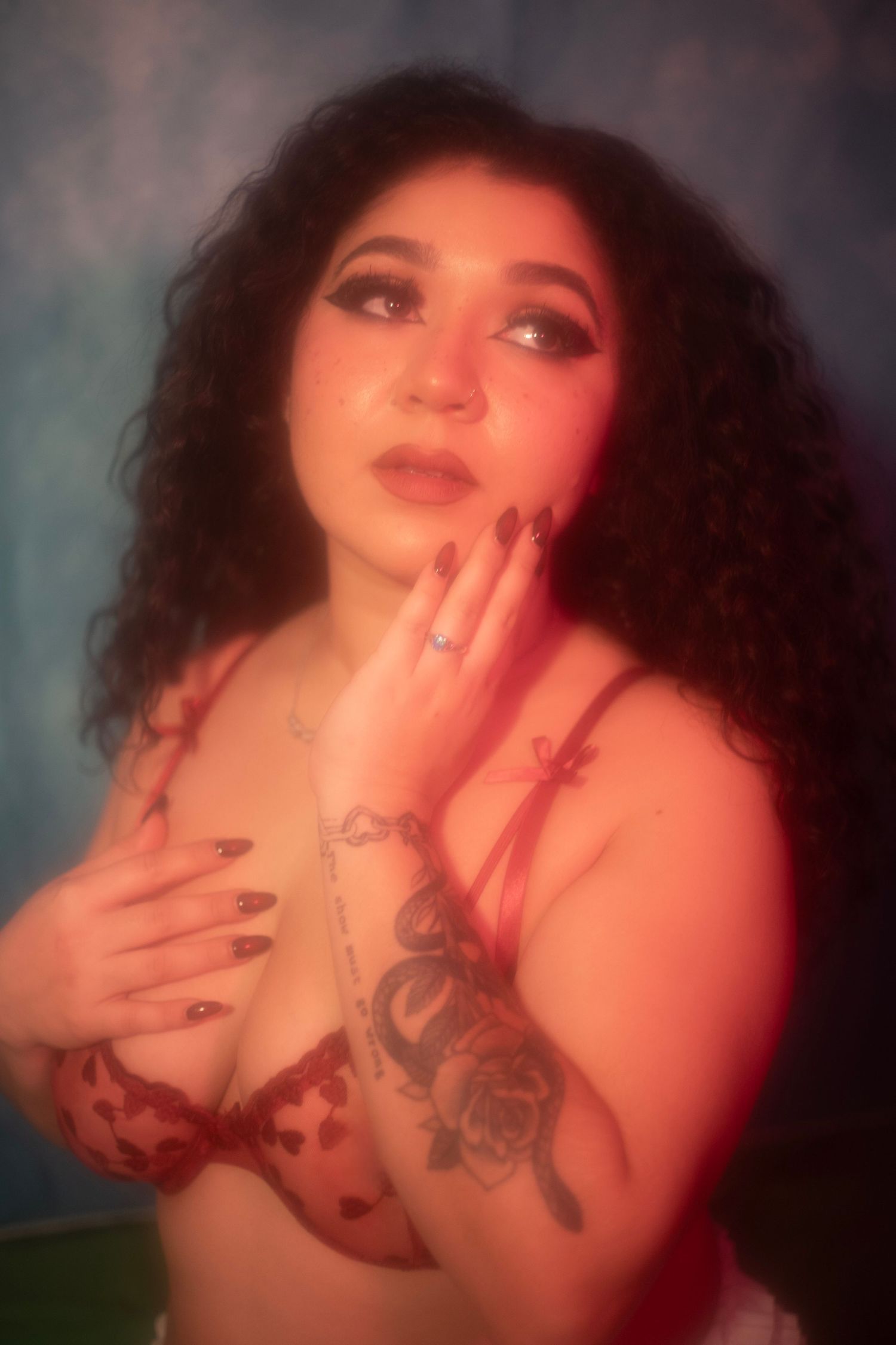 Curly-haired woman in red lace bra with tattoos posing under red lighting against blue background.