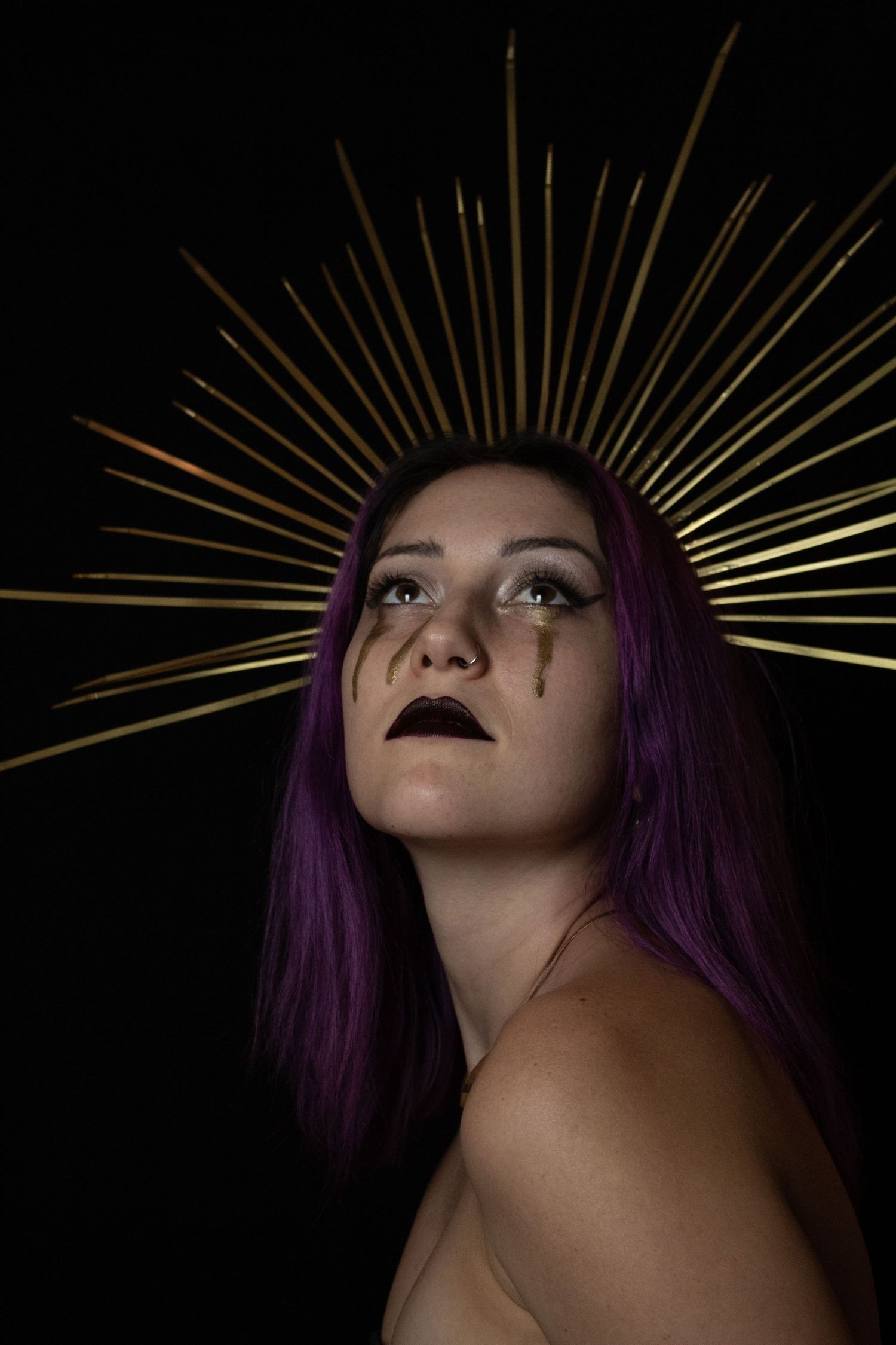 Woman with purple hair and gold sunburst crown headpiece with dramatic gold tear makeup on dark background.