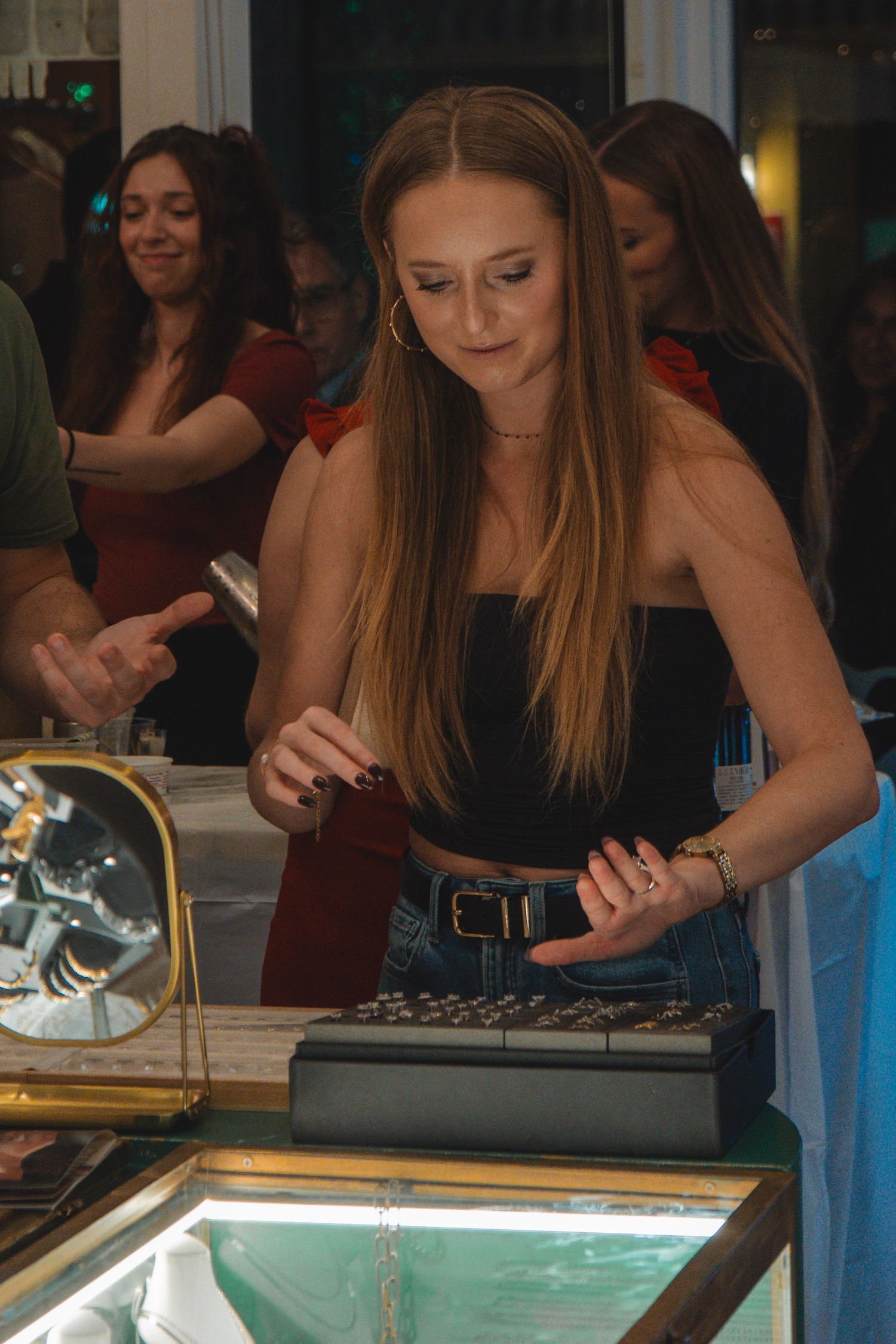 Young woman with long blonde hair browsing jewelry at a display case during an indoor shopping event.