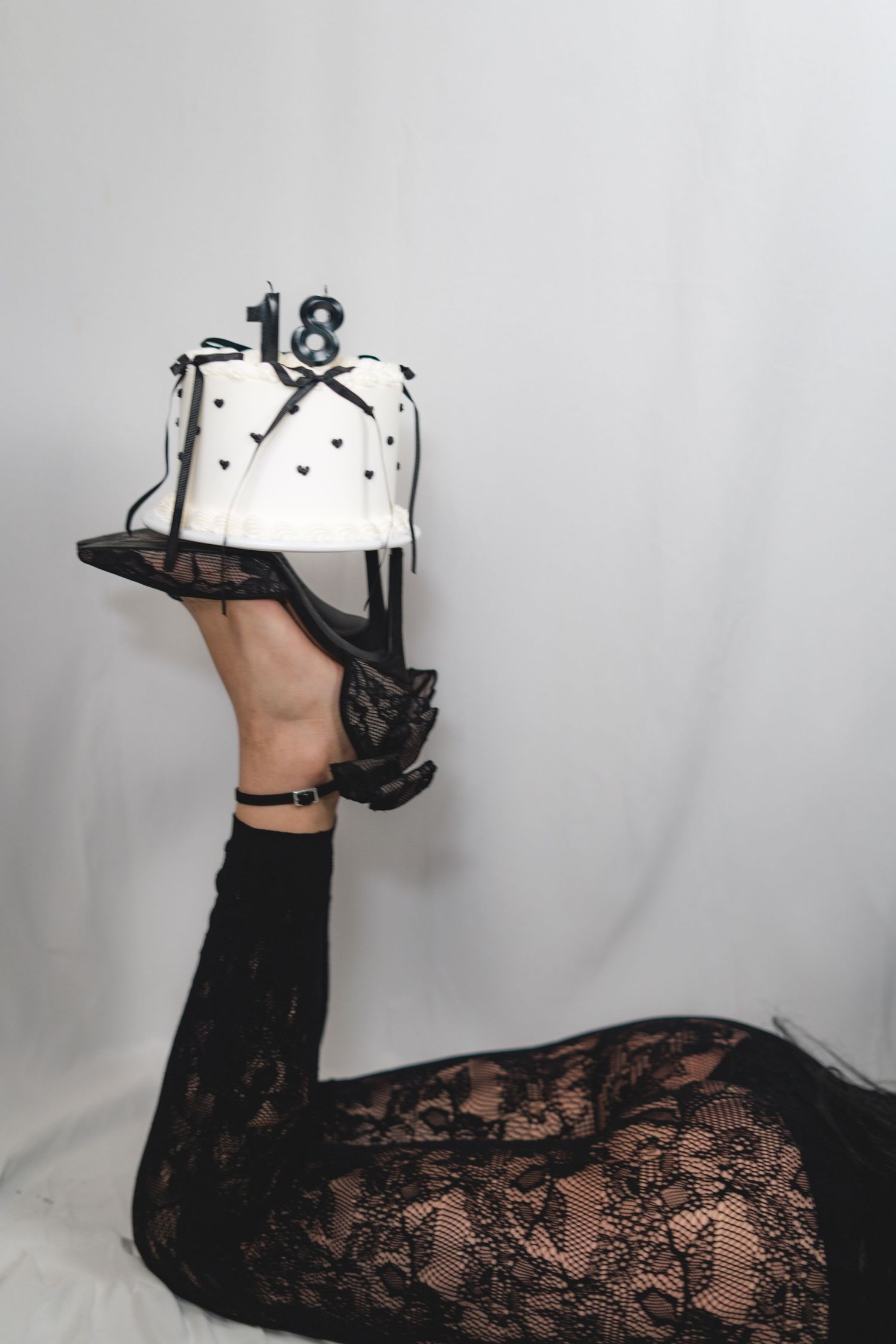 Person in black lace outfit balancing an 18th birthday cake on their raised high-heeled foot.