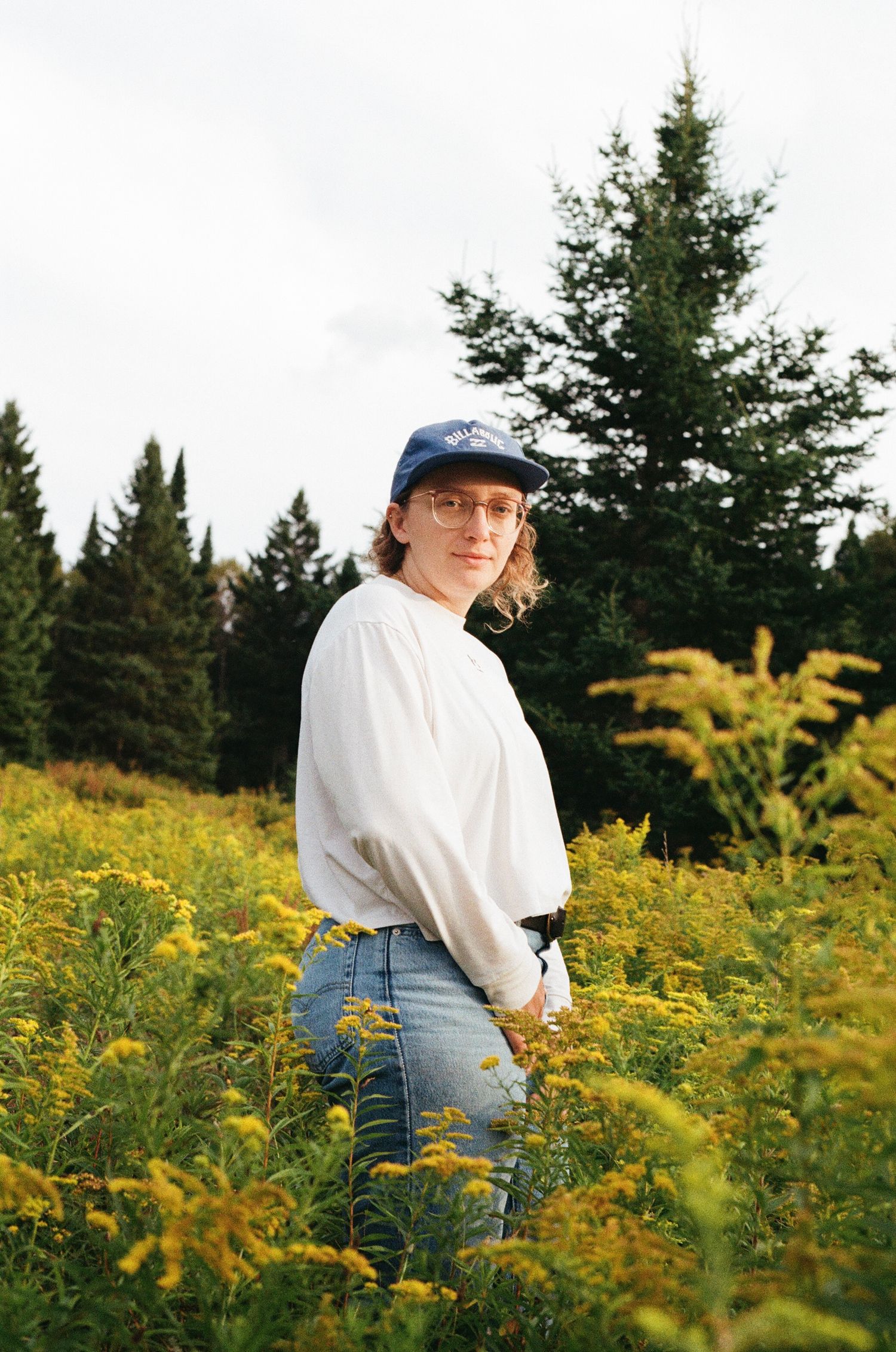A woman wearing glasses, a blue cap, white top, and jeans poses in a field of yellow wildflowers with pine trees behind.