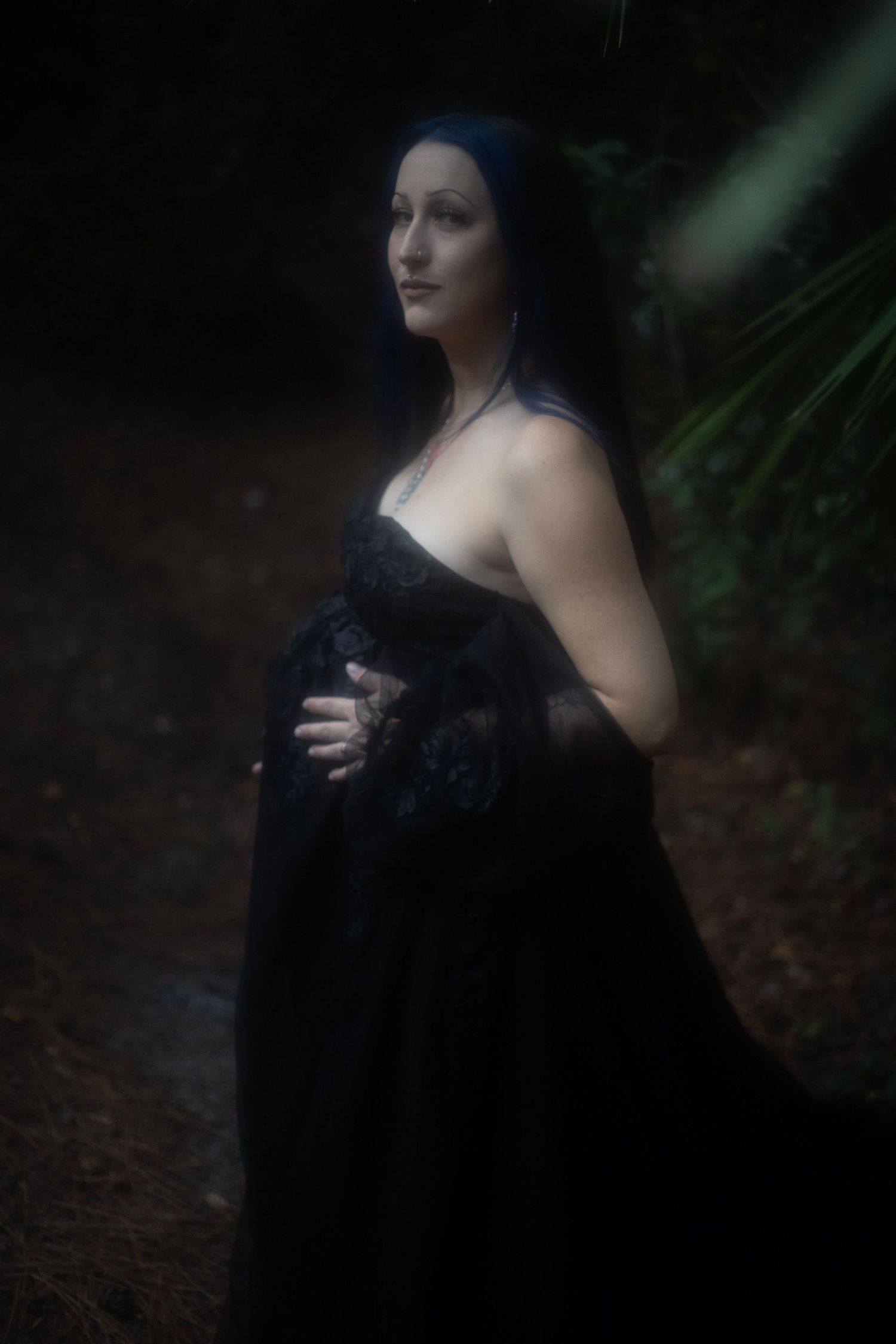 A woman in a black lace gown poses dramatically in a dark forest setting.
