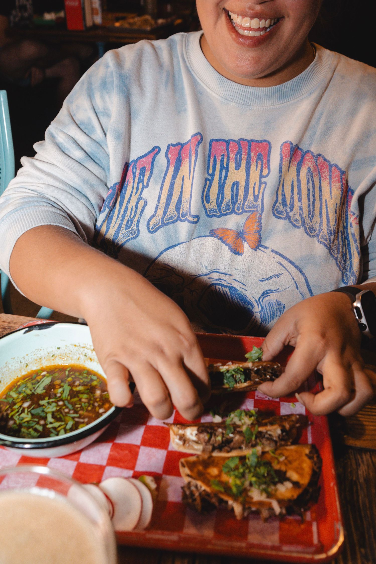 A smiling person enjoying tacos on a red checkered tray with a bowl of salsa at a casual restaurant.
