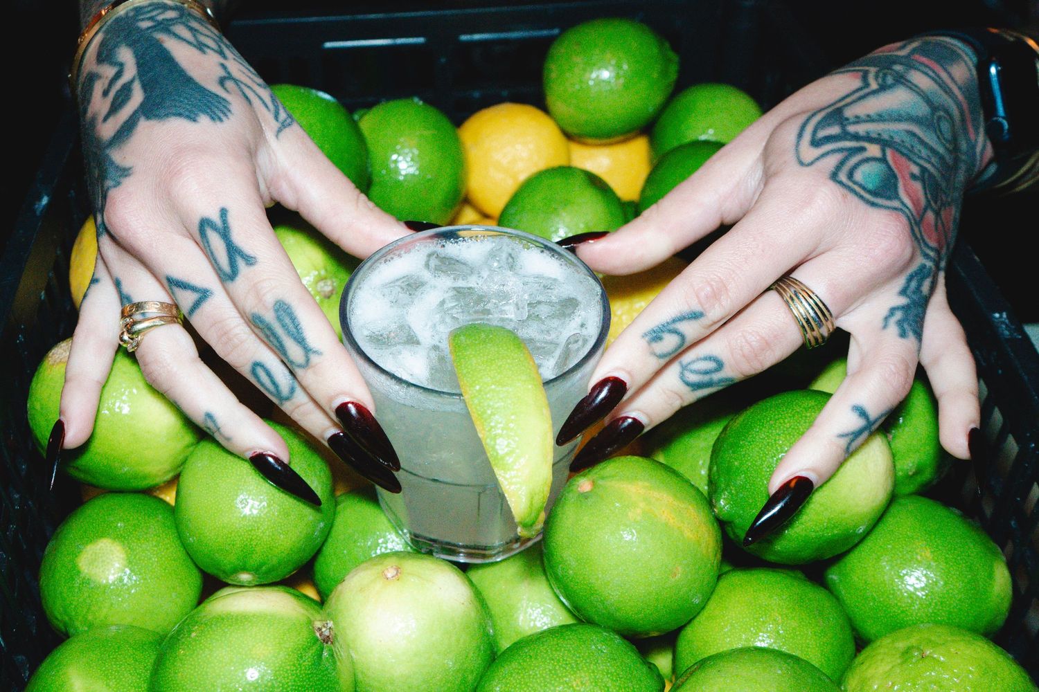 Tattooed hands with black nails holding a lime margarita cocktail surrounded by fresh limes.