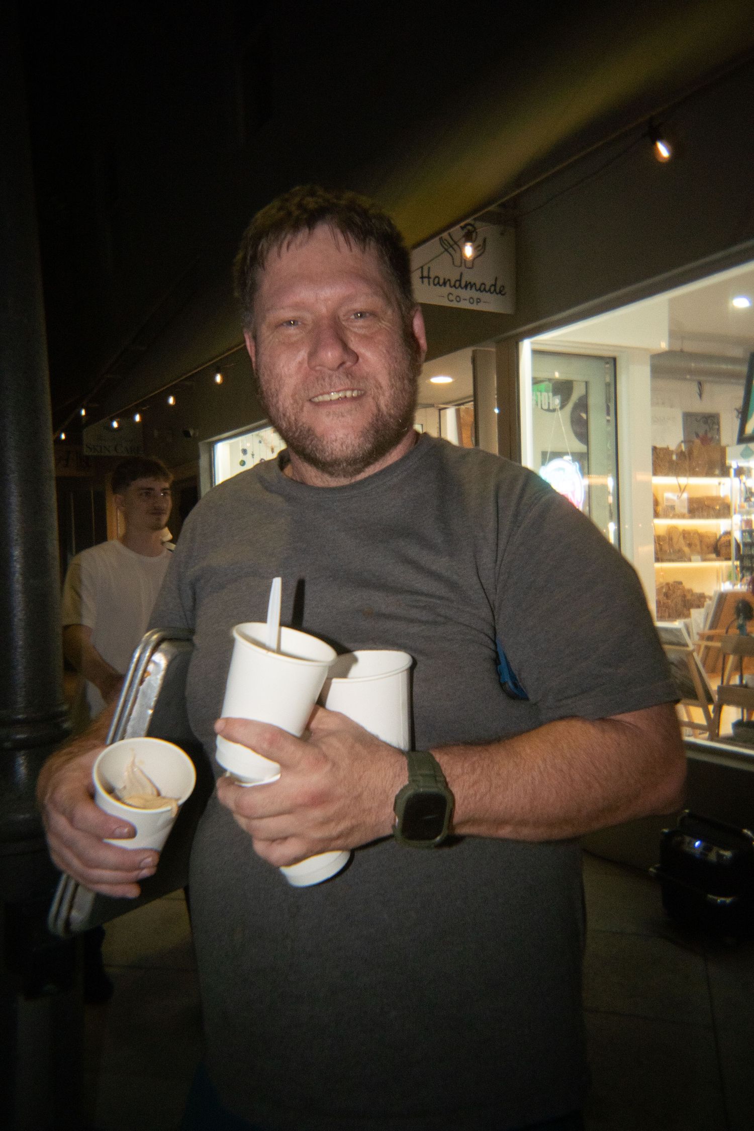 Man in gray t-shirt holding multiple white paper cups and straws outside a shop at night.