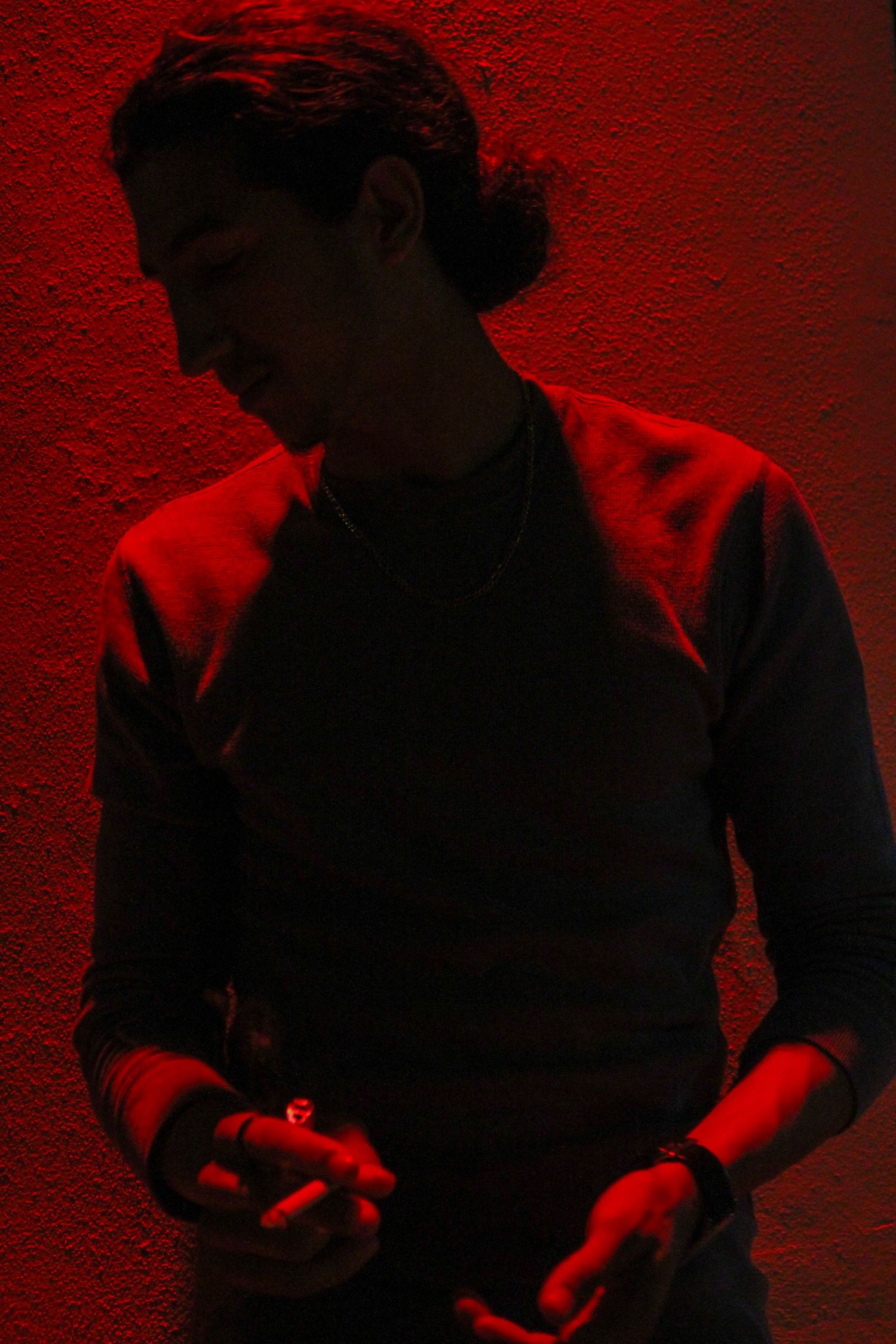 Man in dark sweater poses under dramatic red lighting holding a cigarette.
