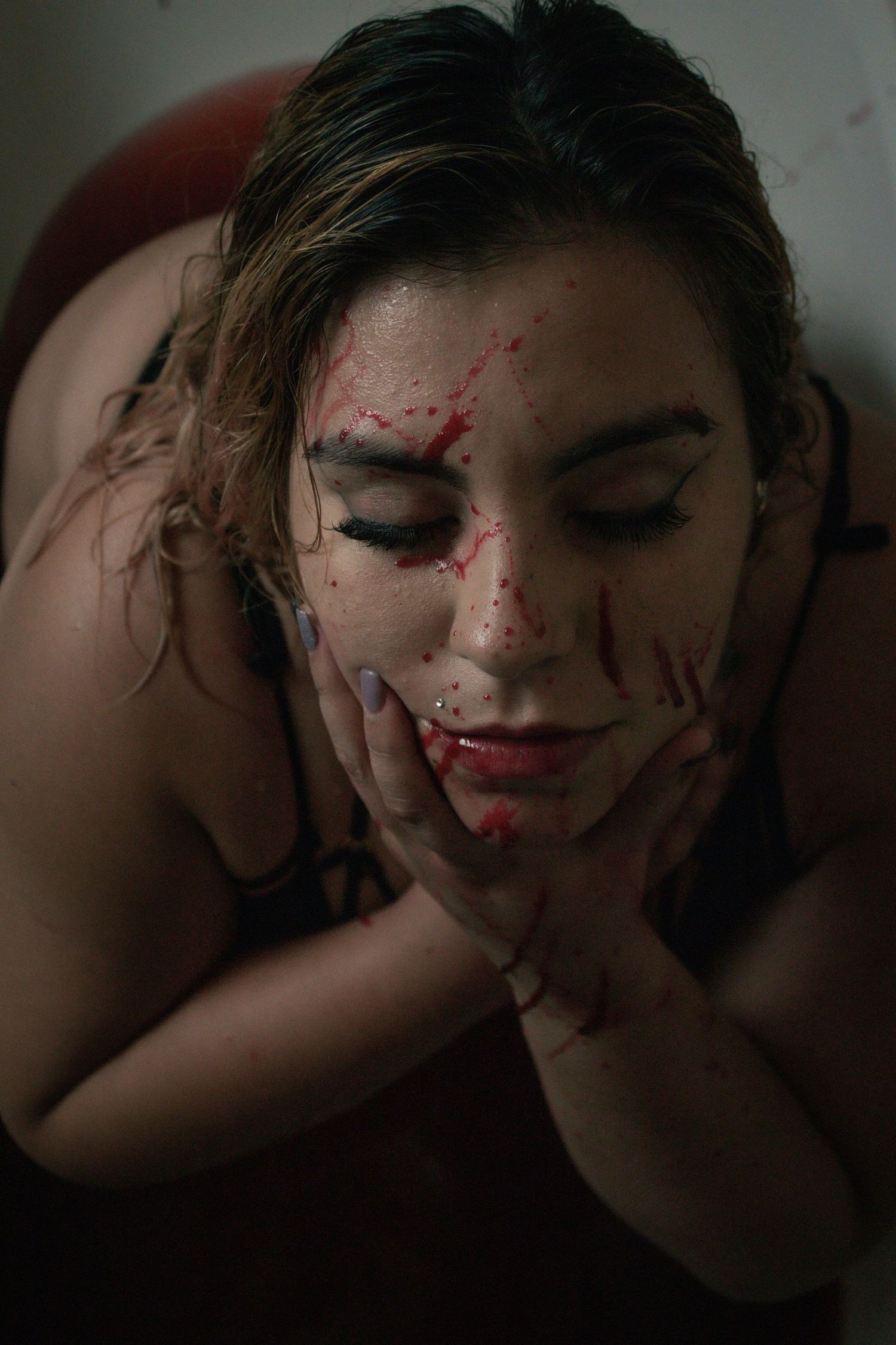 Woman with eyes closed and red paint splattered across her face holding her hands to her cheeks.