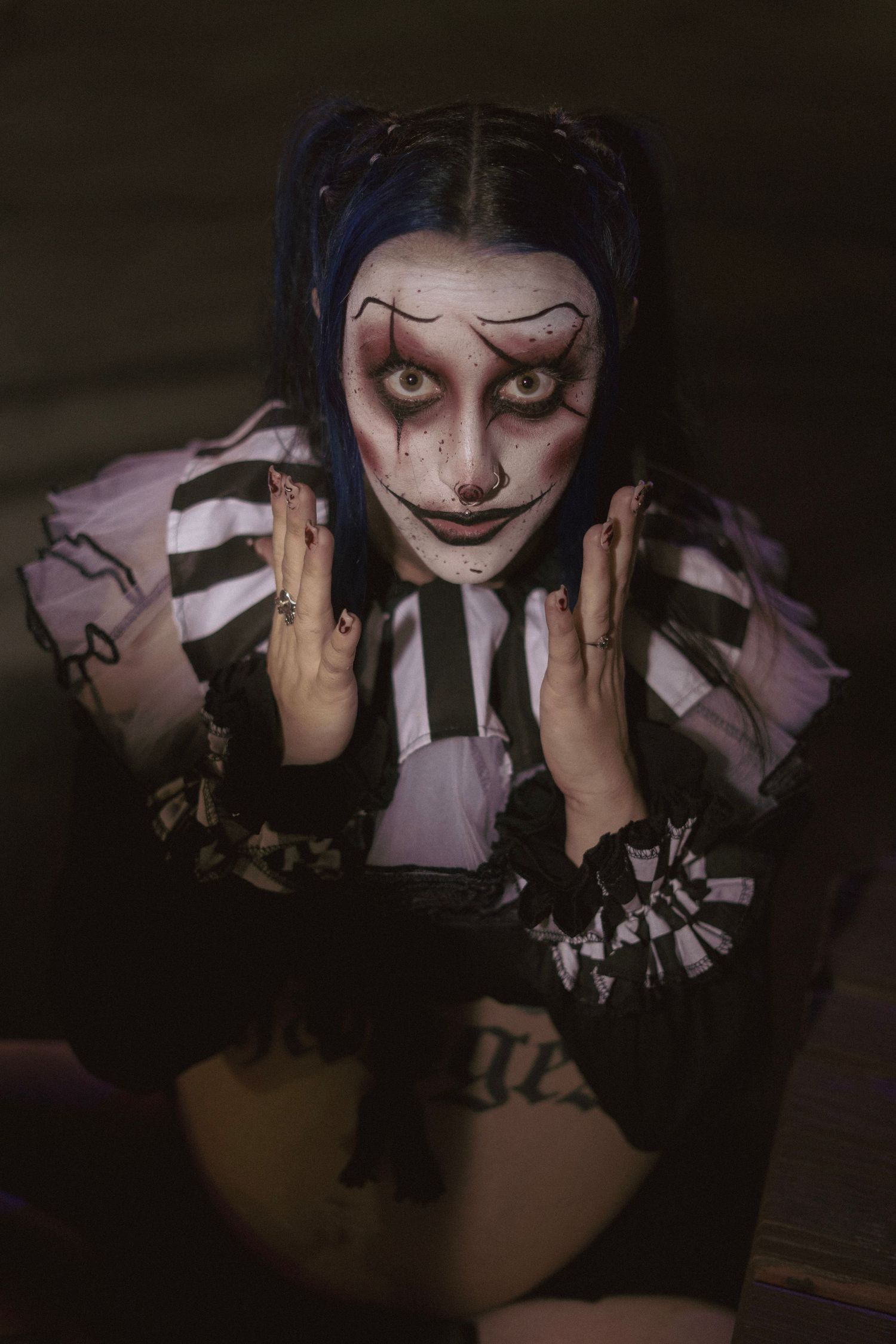 Woman in dramatic clown makeup with blue pigtails and black-and-white costume poses with hands raised in dark setting.